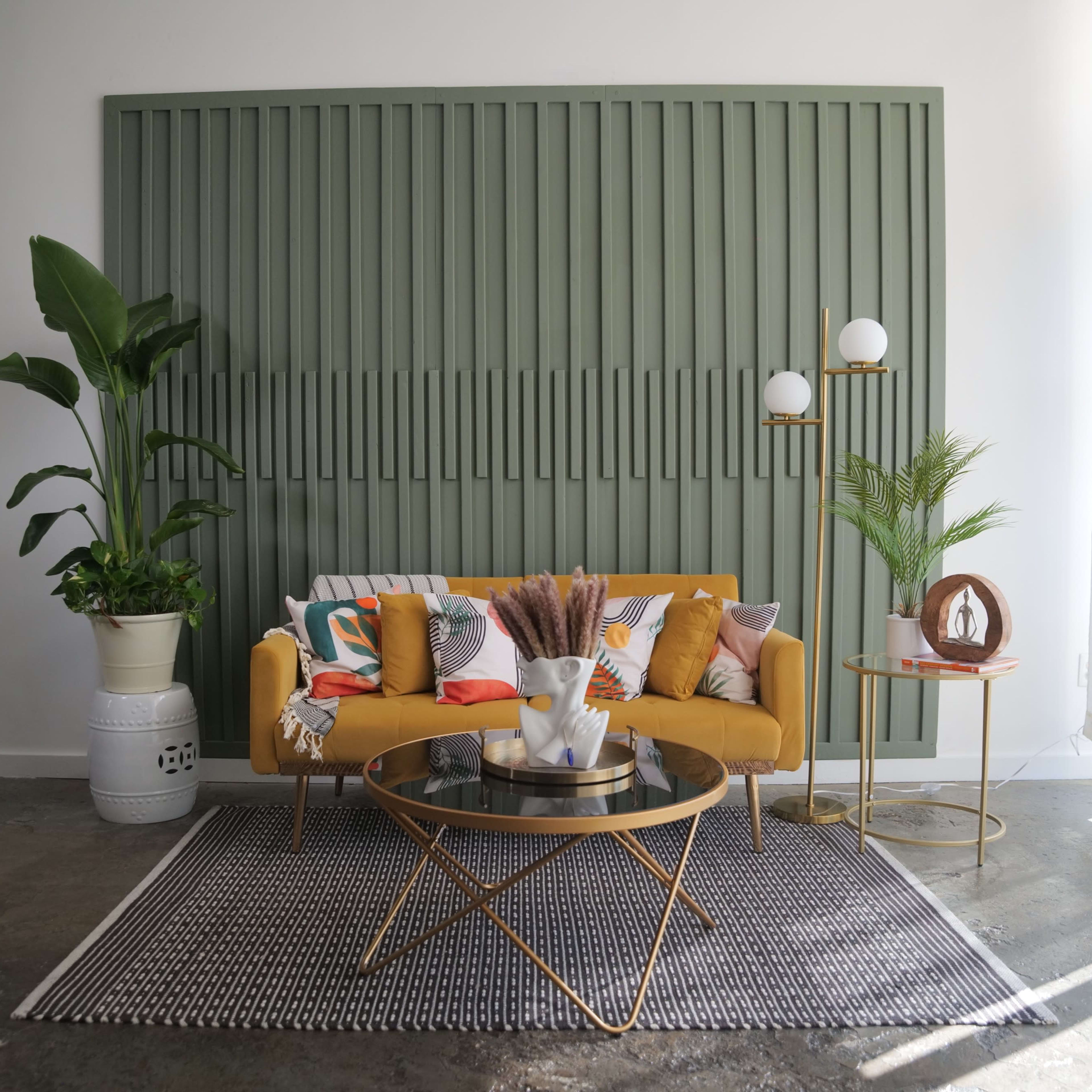 A modern living room features a mustard-colored sofa with decorative pillows, a round coffee table, and potted plants against a green paneled wall.