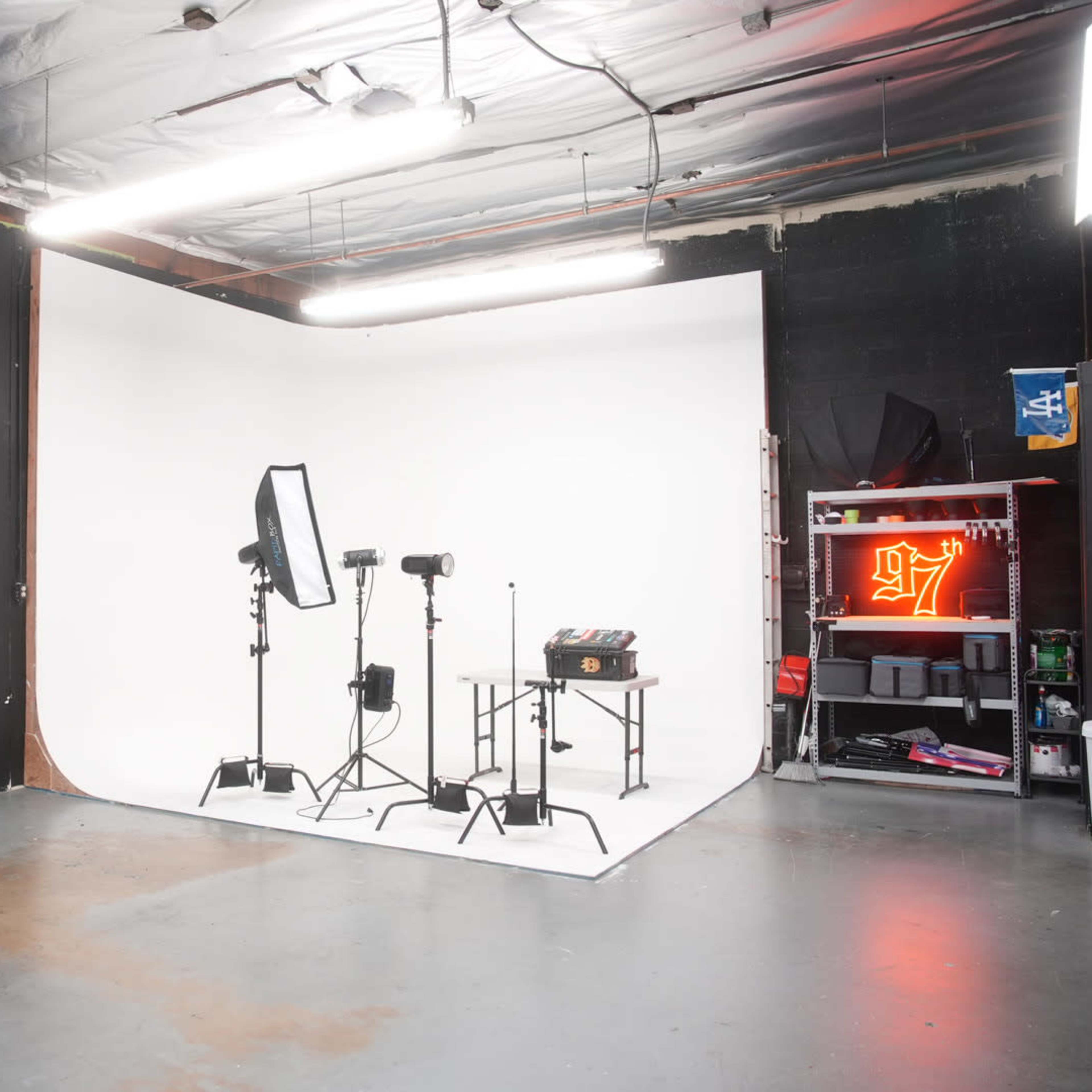 A photography studio featuring a white backdrop, lighting equipment, a table, and a storage area with various items.