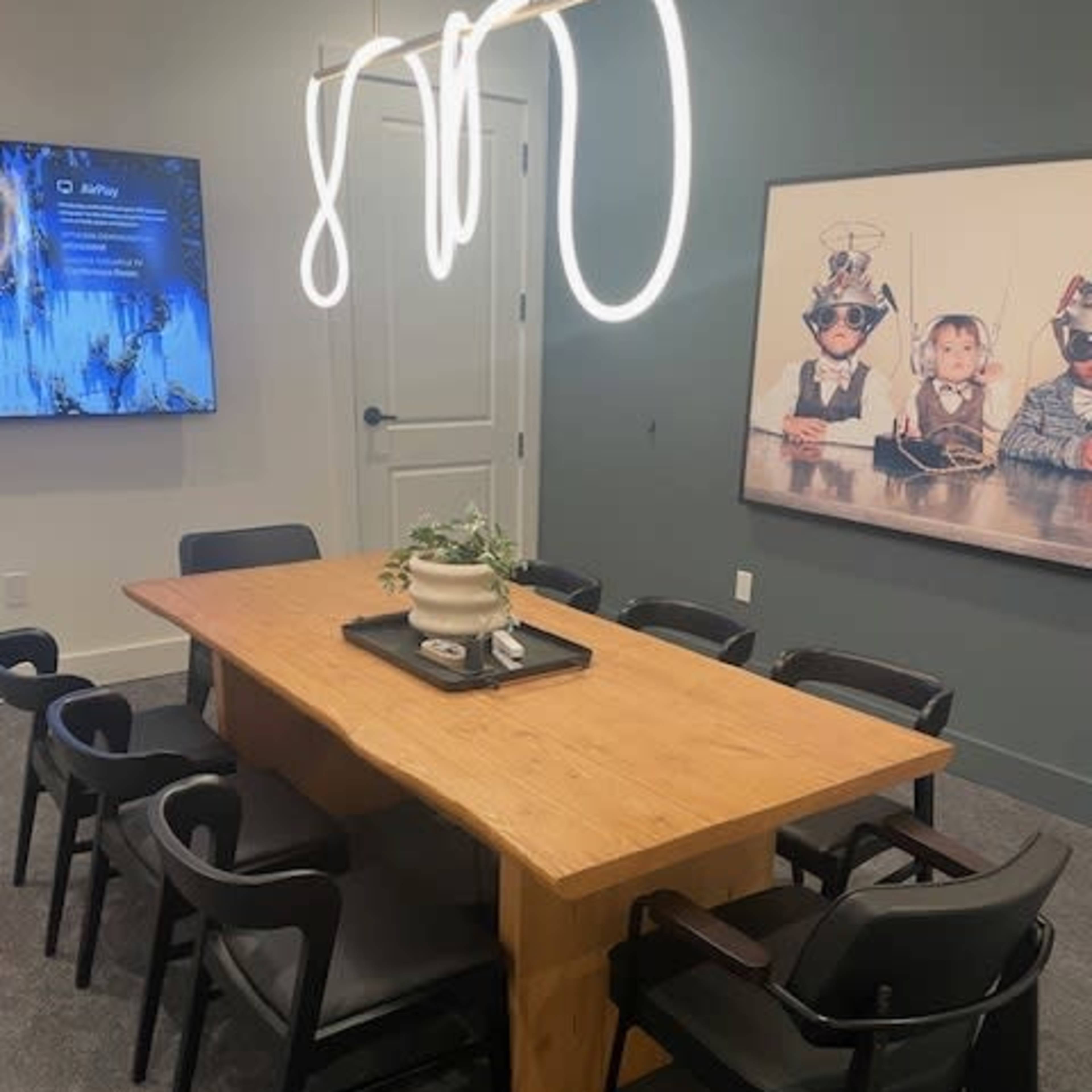 A modern conference room with a large wooden table surrounded by black chairs, a decorative plant on the table, and a wall featuring a framed photograph of children in playful costumes.
