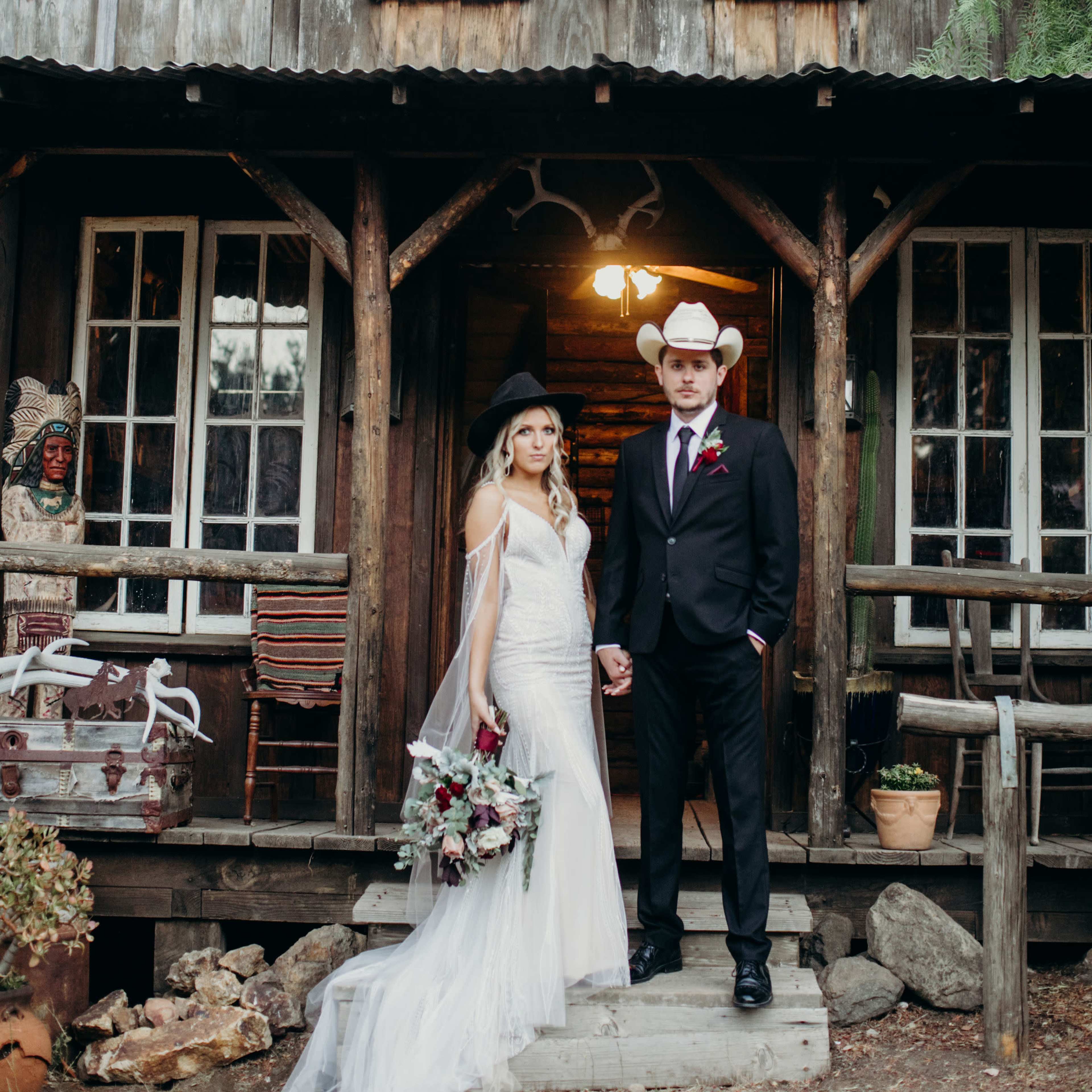 A bride and groom stand together in front of a rustic wooden cabin, holding hands and dressed elegantly for their wedding.