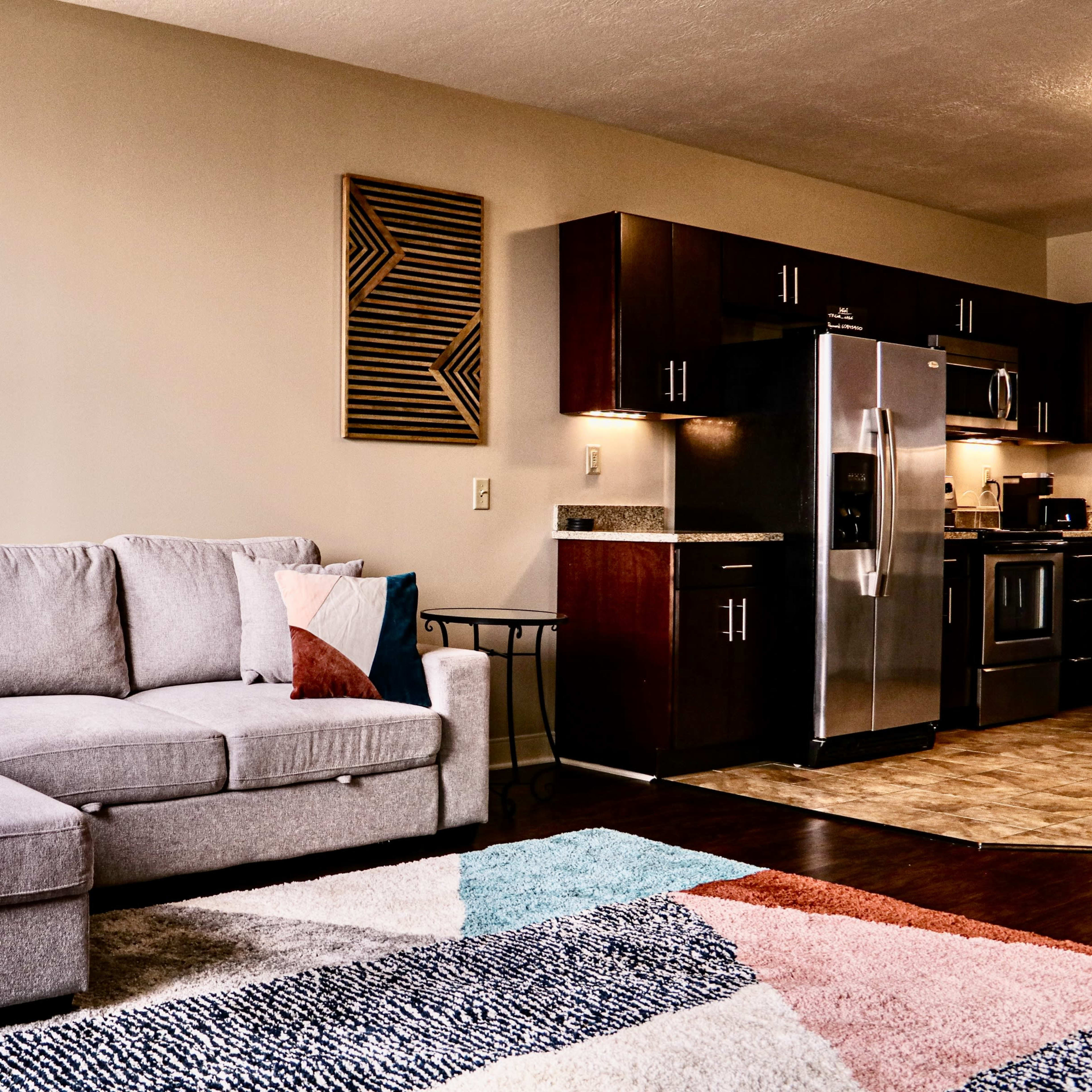 A modern living room featuring a gray sectional sofa, a multi-colored area rug, and a kitchen with dark wooden cabinets and stainless steel appliances.