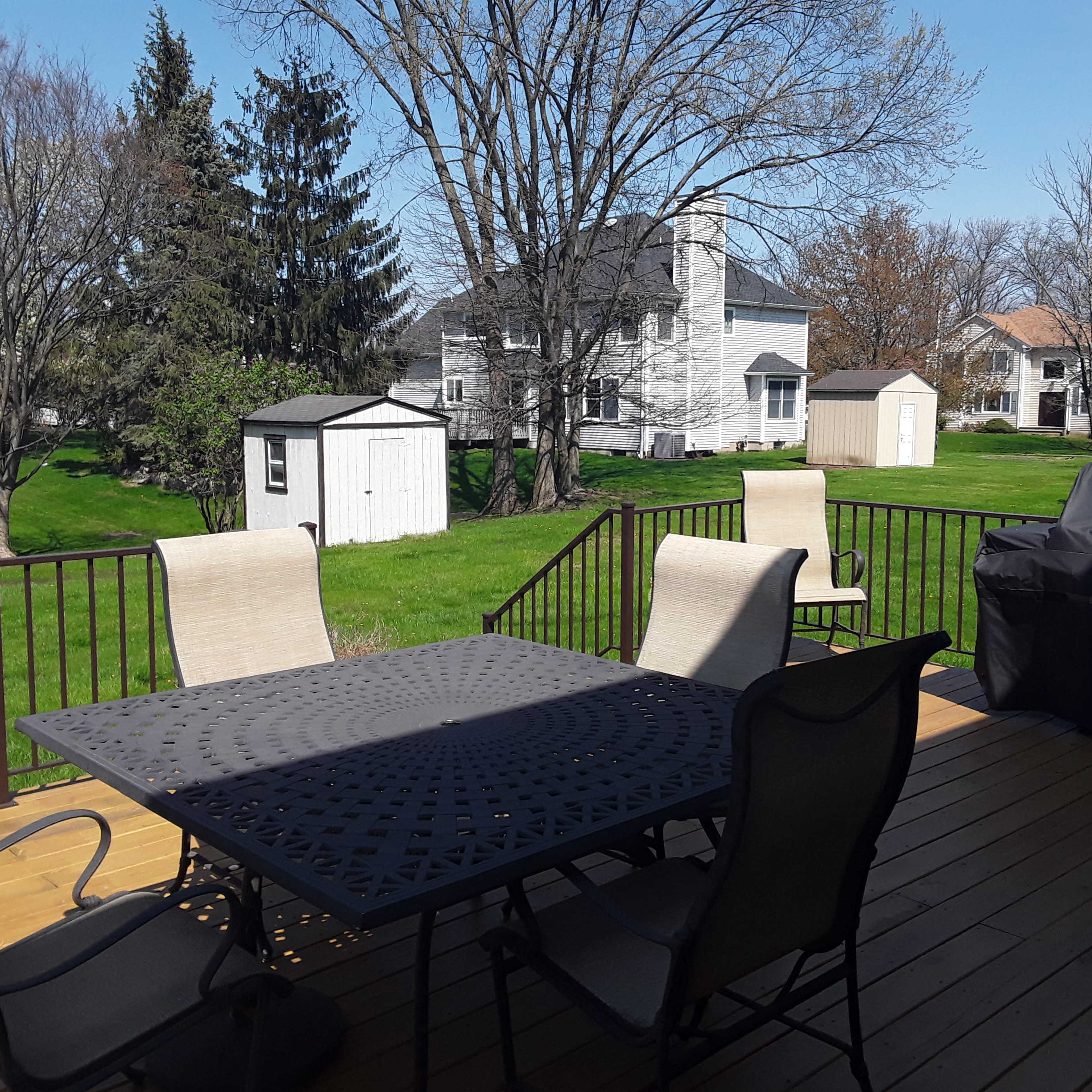 A deck features a table and chairs, overlooking a grassy area with a shed and several houses in the background.