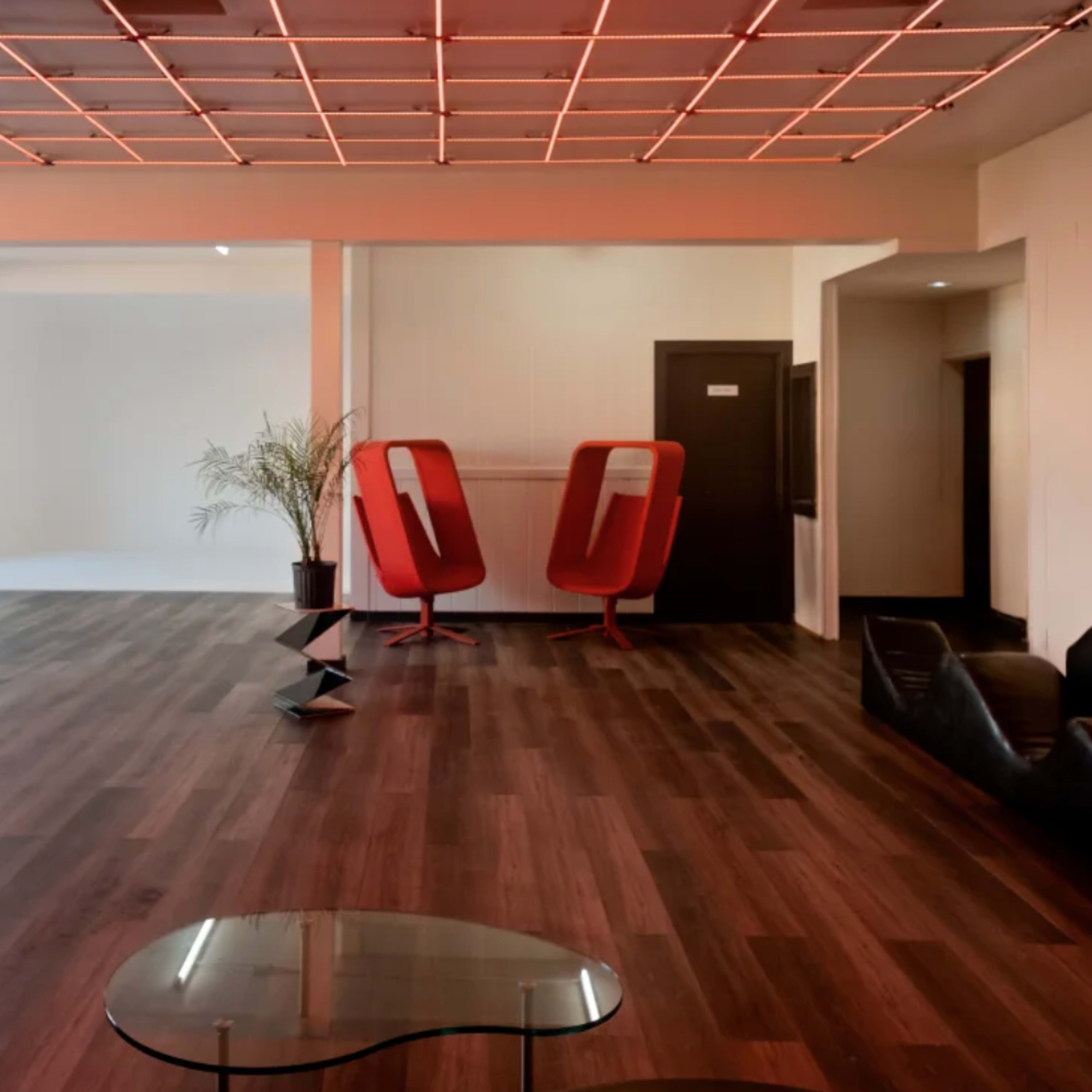 A spacious, modern room with a wooden floor, featuring red seating areas, two red chairs, a hanging chair, and a geometric black sculpture.