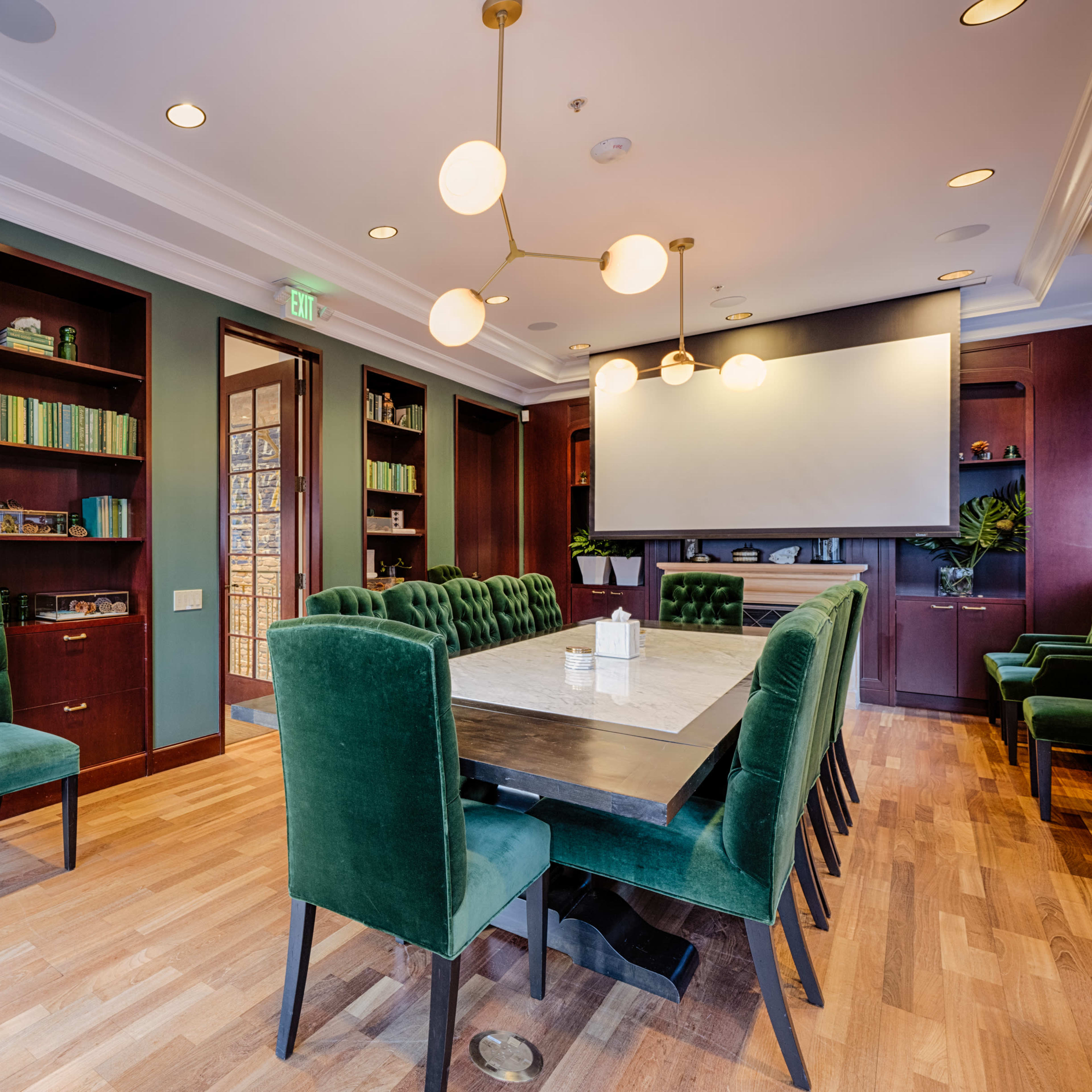 A conference room features a large table surrounded by green upholstered chairs, with shelves of books and a projector screen on the wall.