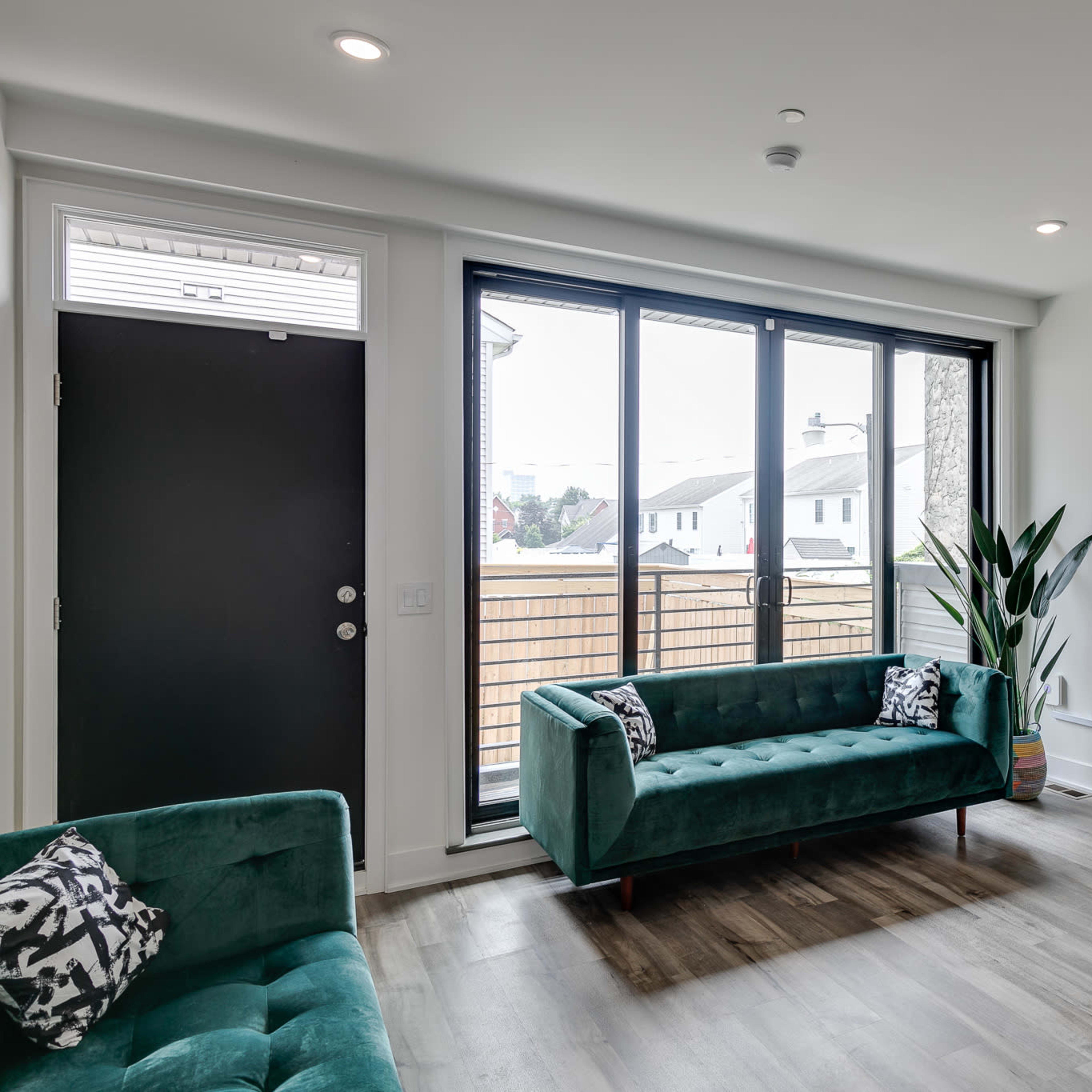 A modern living room with two green sofas, a television mounted on the wall, a wooden shelf with decorative items, and a large window overlooking an outdoor space.