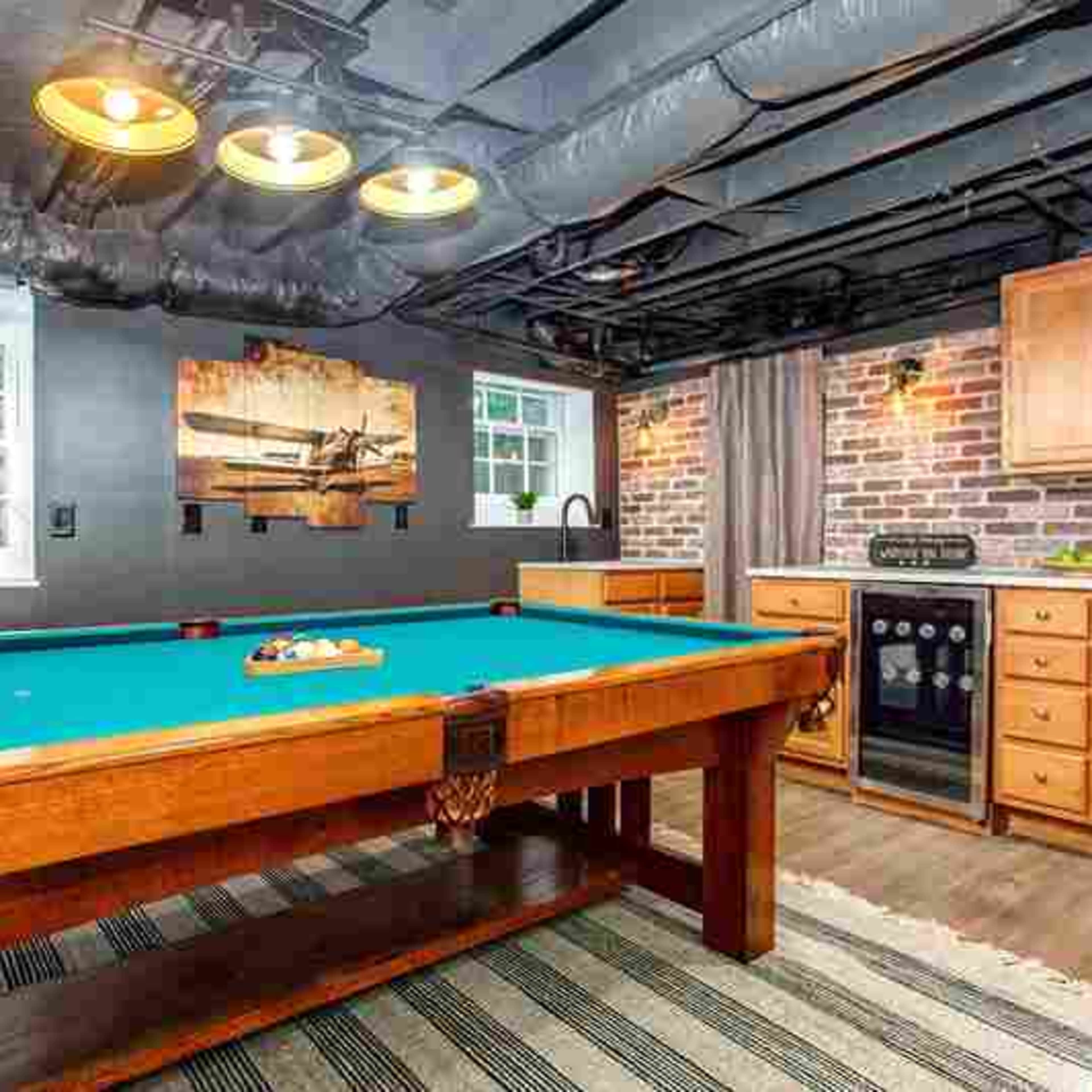 A basement game room with a pool table, exposed brick walls, a kitchenette, and overhead lighting.