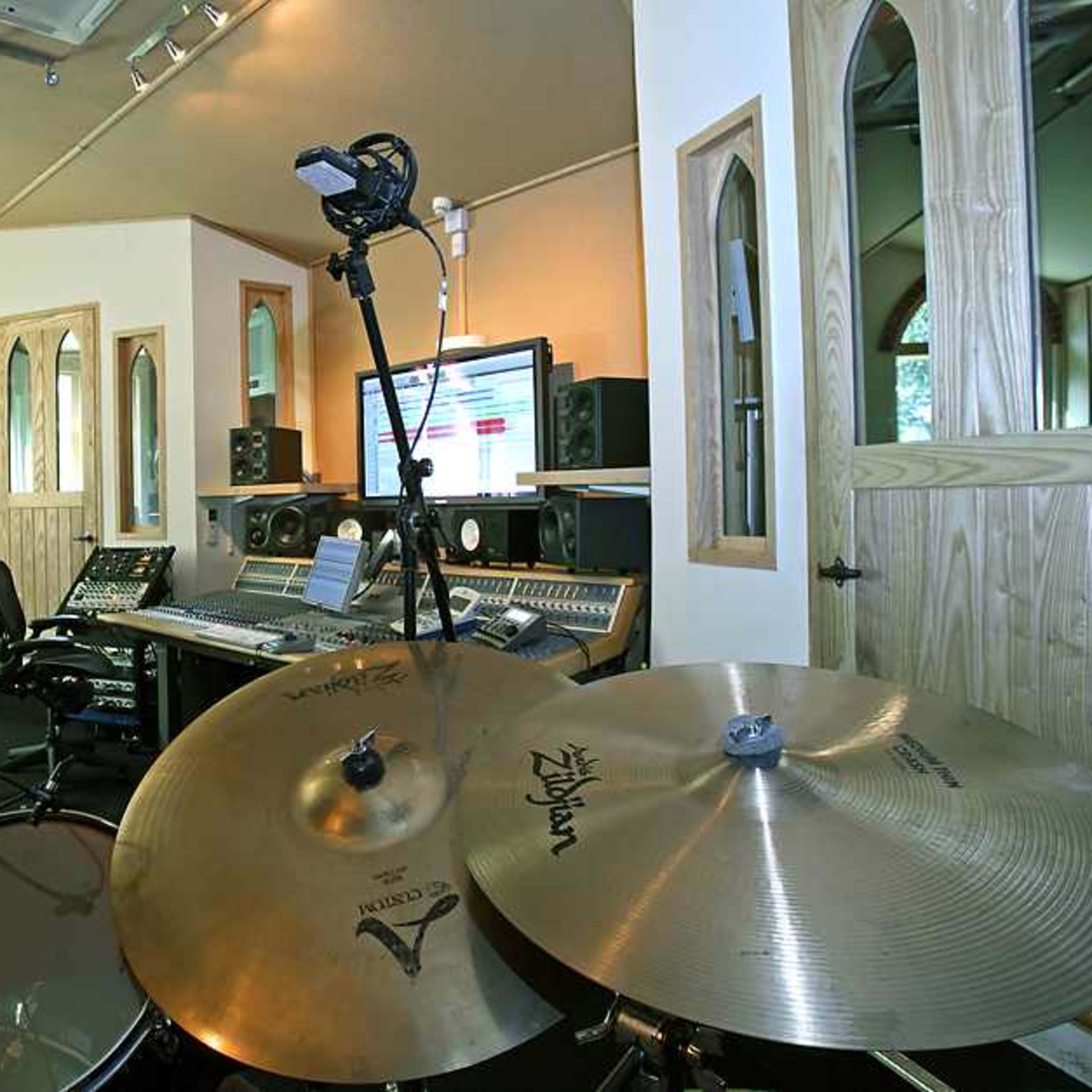 A recording studio featuring a drum set in the foreground, with microphones set up and a control desk in the background surrounded by audio equipment.