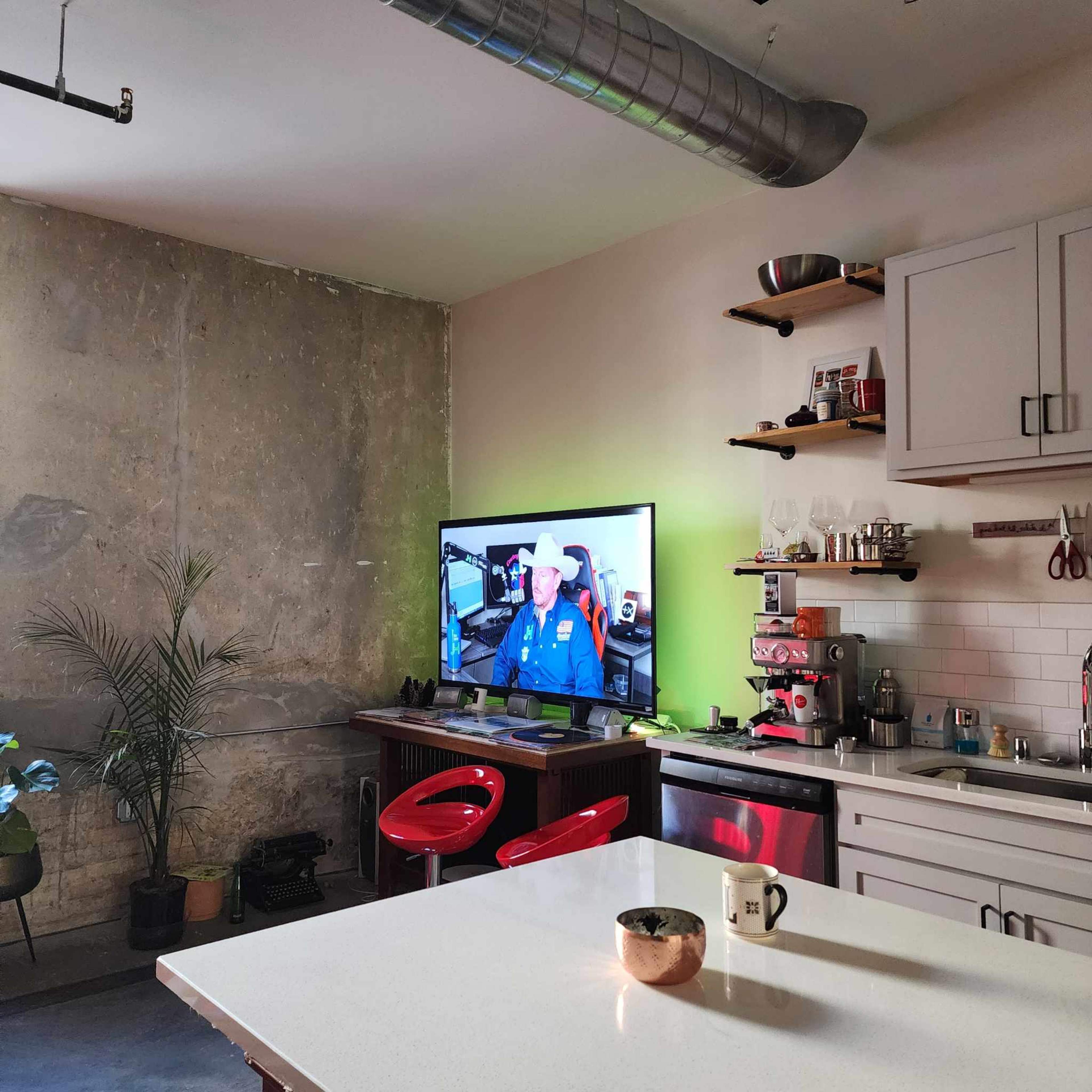 A modern kitchen features a green accent wall, industrial elements, a large TV displaying a man in a cowboy hat, and a bar area with red stools.