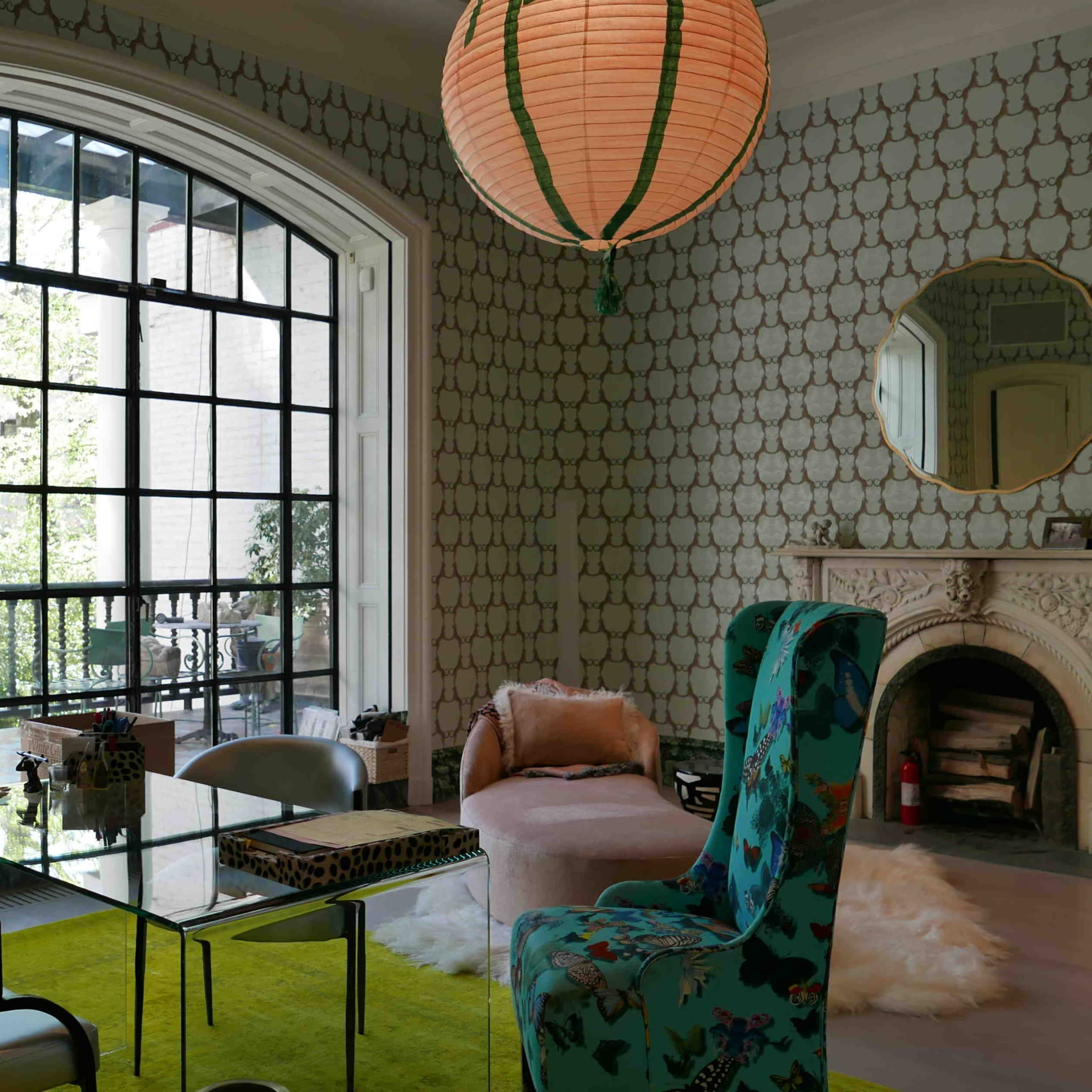 A modern living room features a glass table, a patterned wall, a large window overlooking greenery, and eclectic seating arrangements.