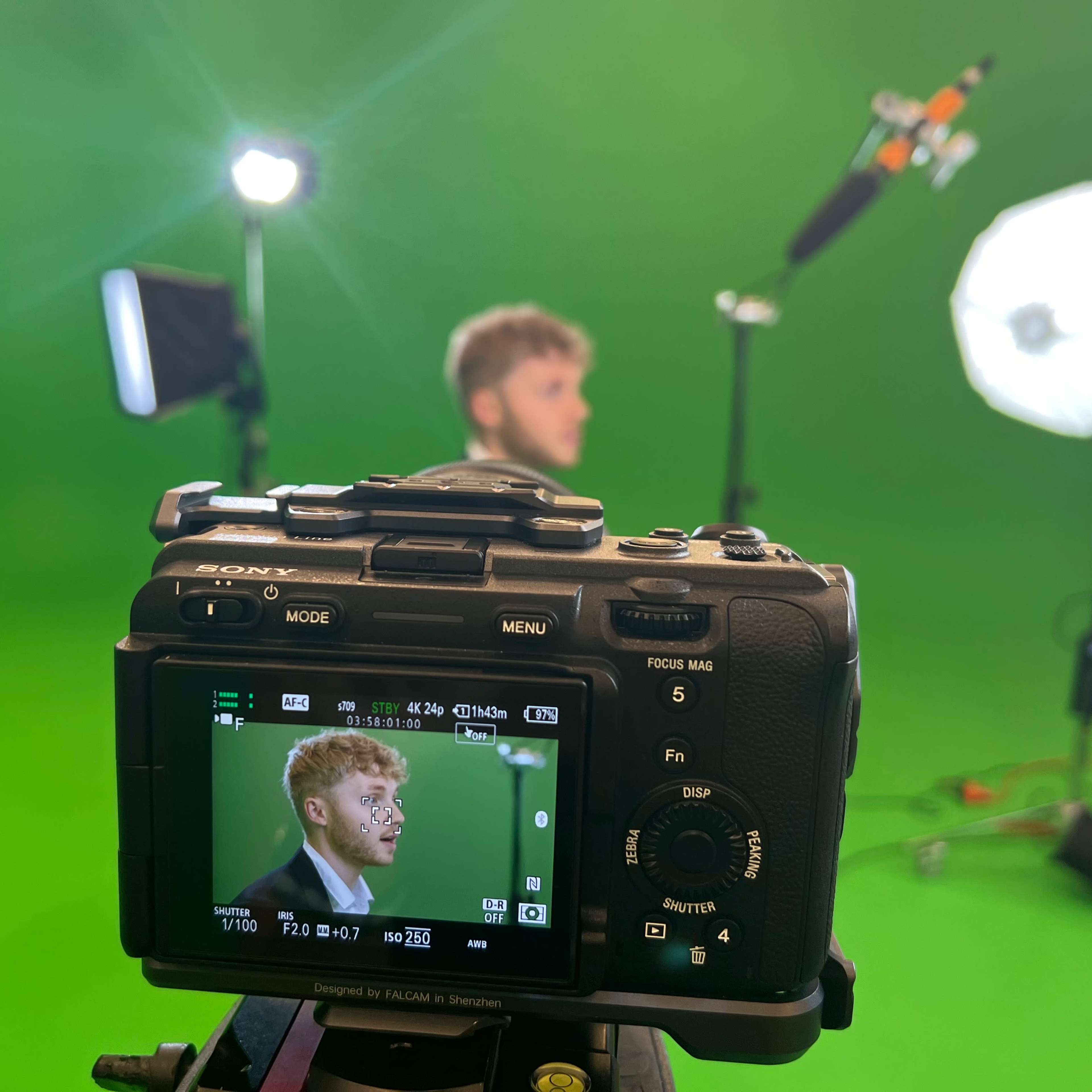 A camera is positioned on a tripod with its screen displaying a man in front of a green screen, while studio lights illuminate the scene.