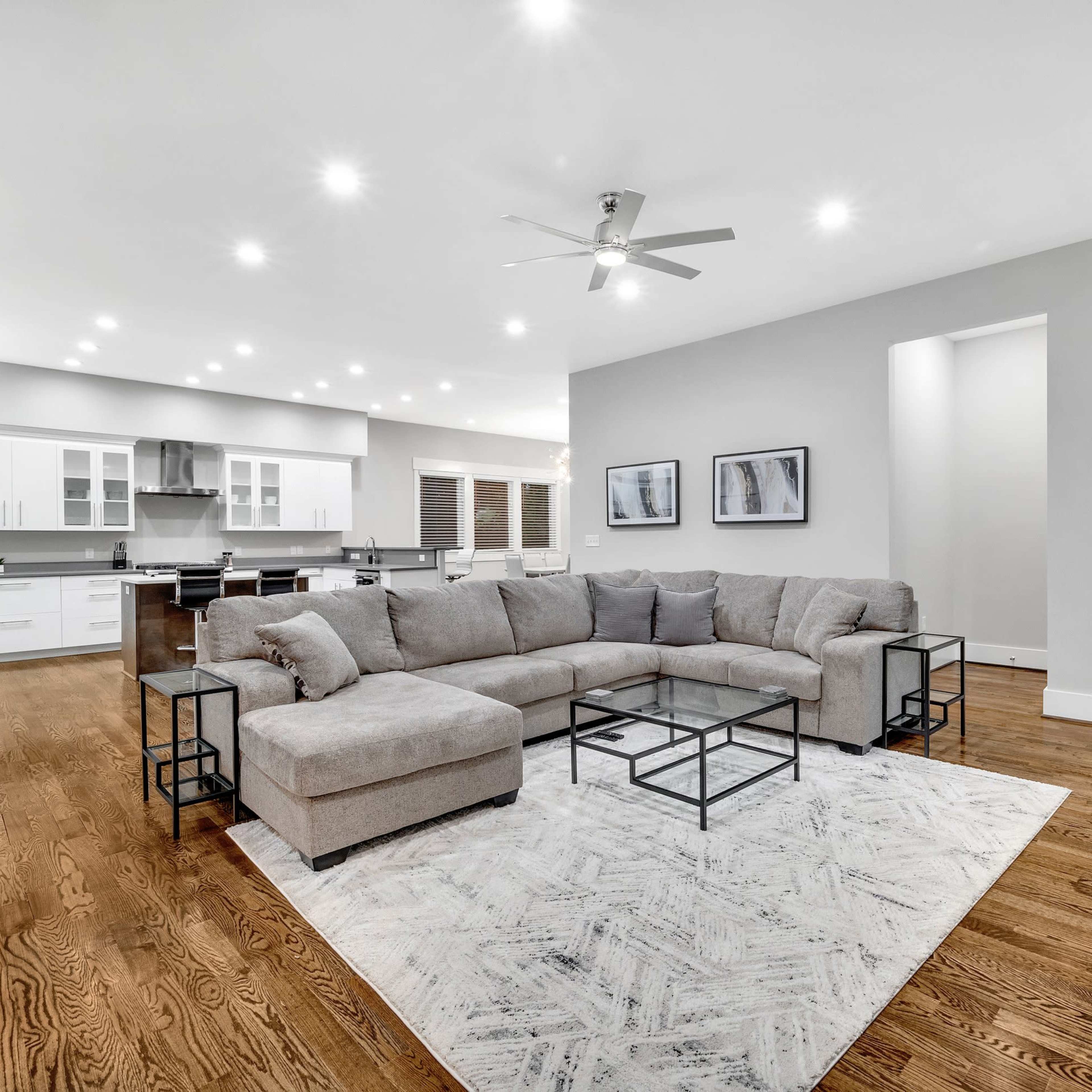 A spacious living room features a gray sectional sofa, black coffee tables, and a large area rug on polished hardwood floors, with a modern kitchen visible in the background.