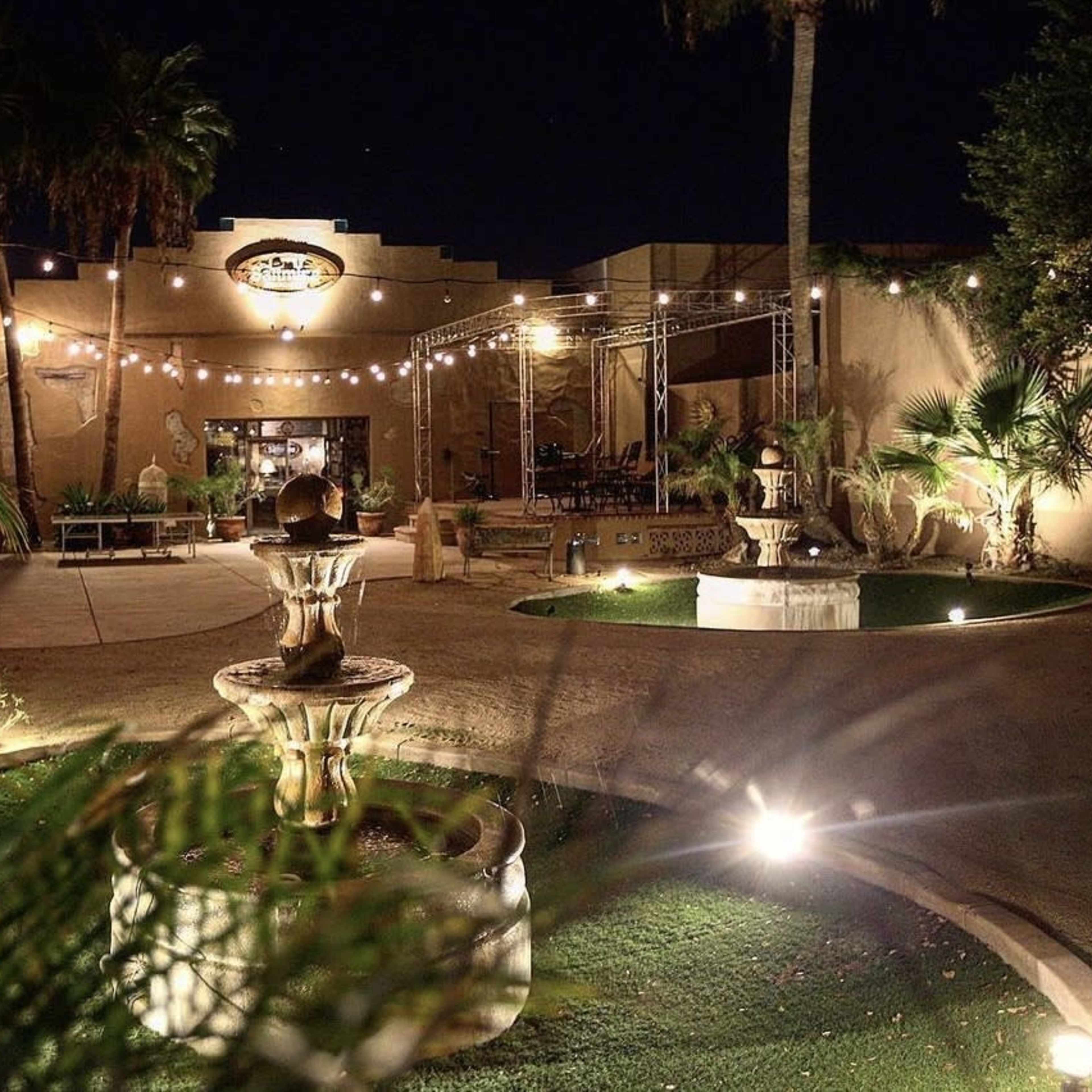 A well-lit outdoor courtyard at night, featuring a fountain surrounded by palm trees and string lights above.