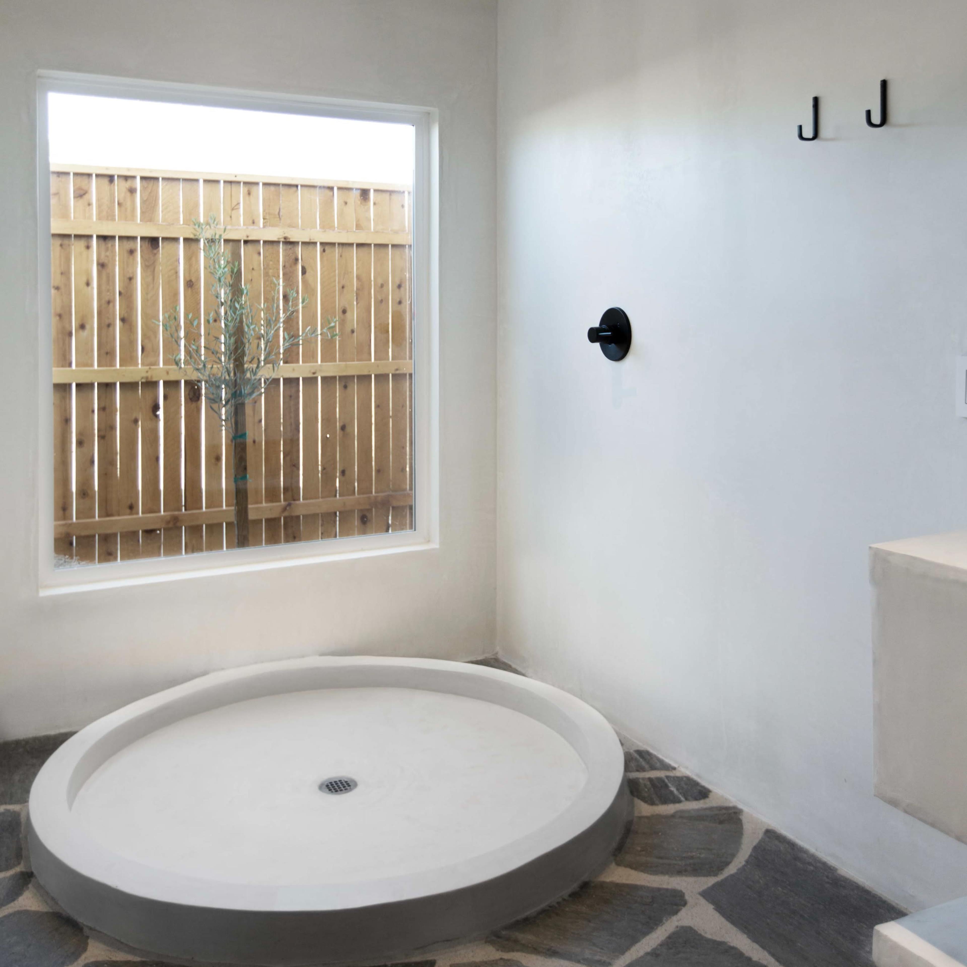 A minimalist bathroom featuring a circular shower base, gray stone flooring, and a window with a view of a wooden fence and a tree.