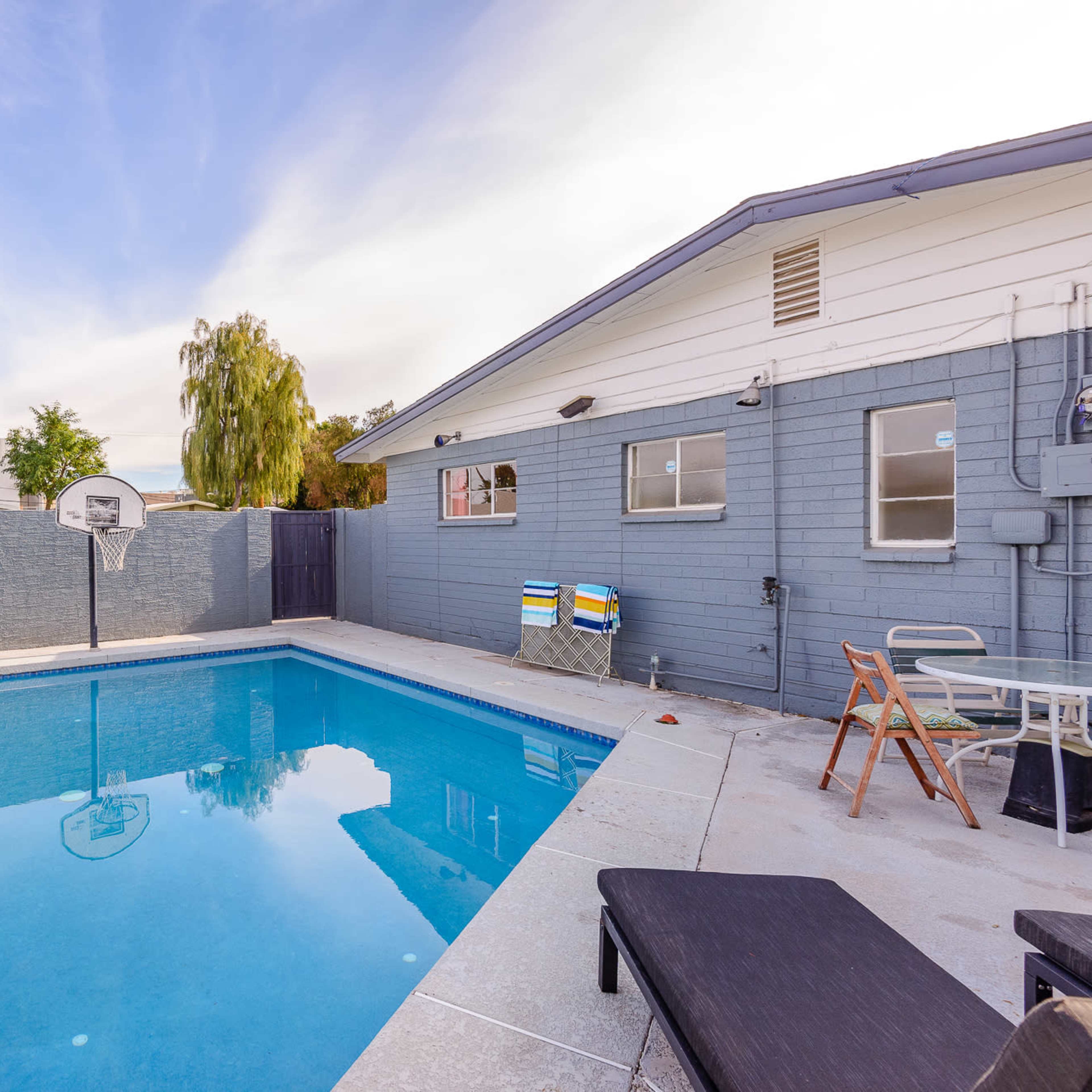 A swimming pool surrounded by a concrete patio features a basketball hoop, beach balls, and a lounge chair.