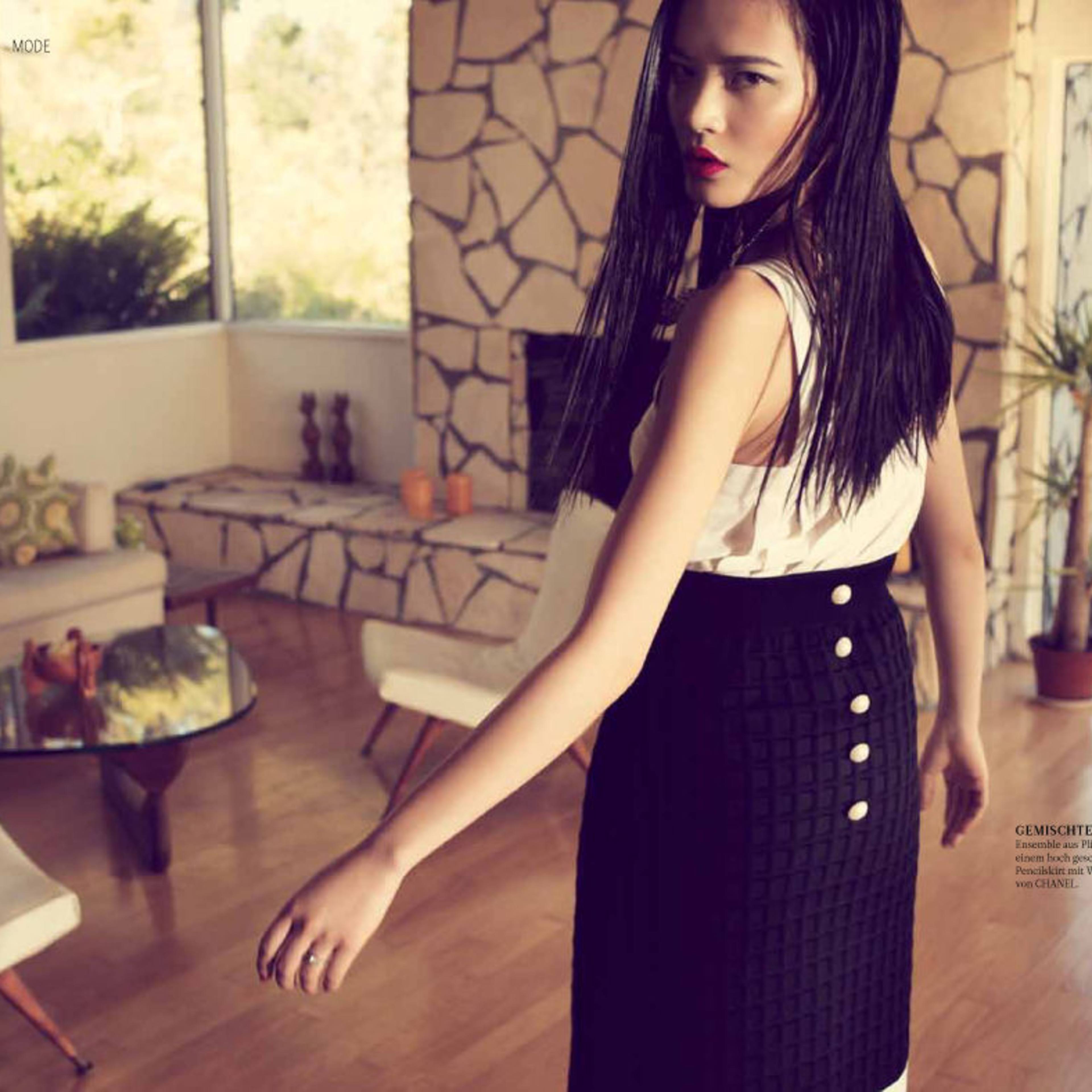 A woman with long hair stands in a spacious, modern room featuring a stone wall and a large window overlooking greenery.