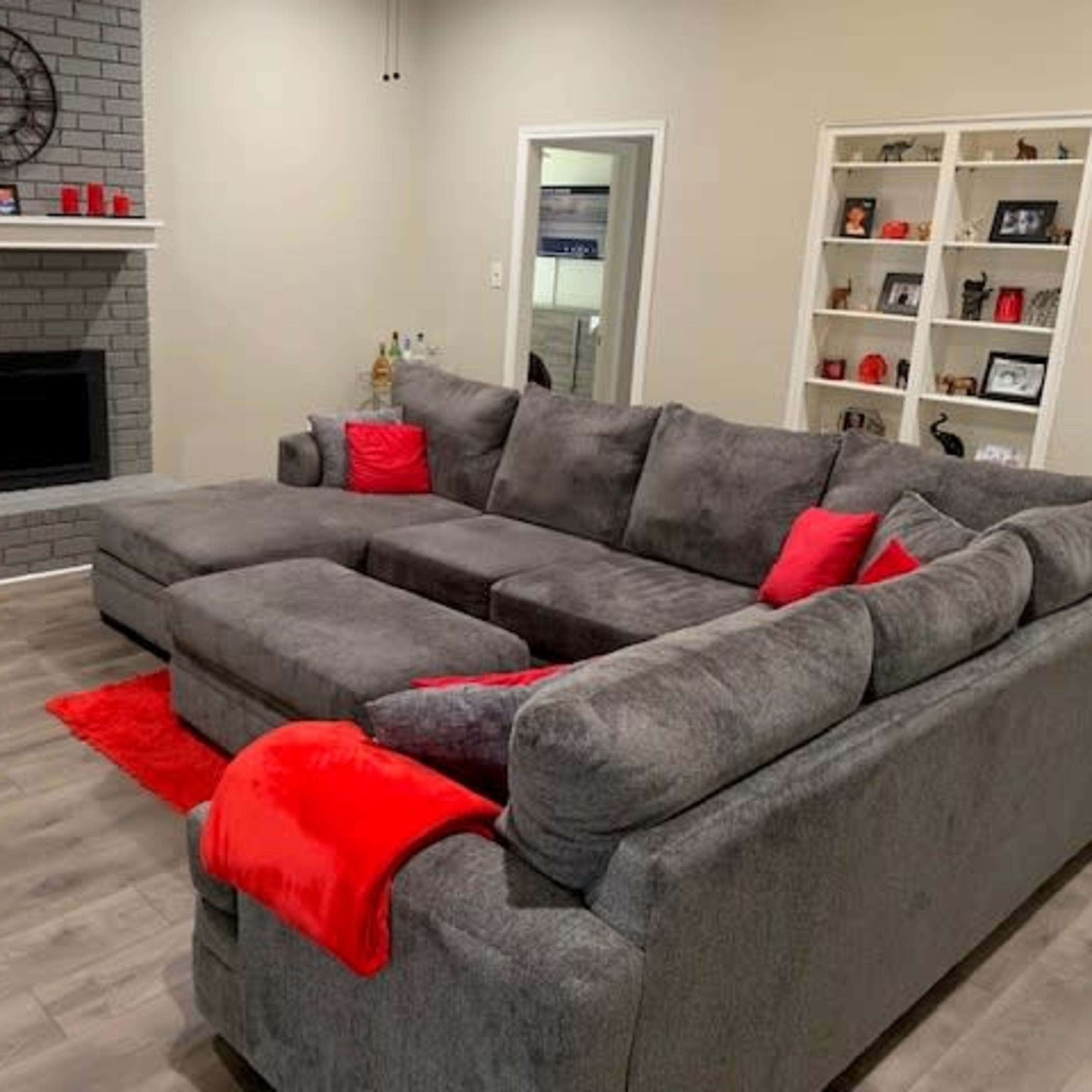 A spacious living room featuring a gray sectional sofa with red accent pillows, a brick fireplace, and shelves displaying framed photos and decorative items.