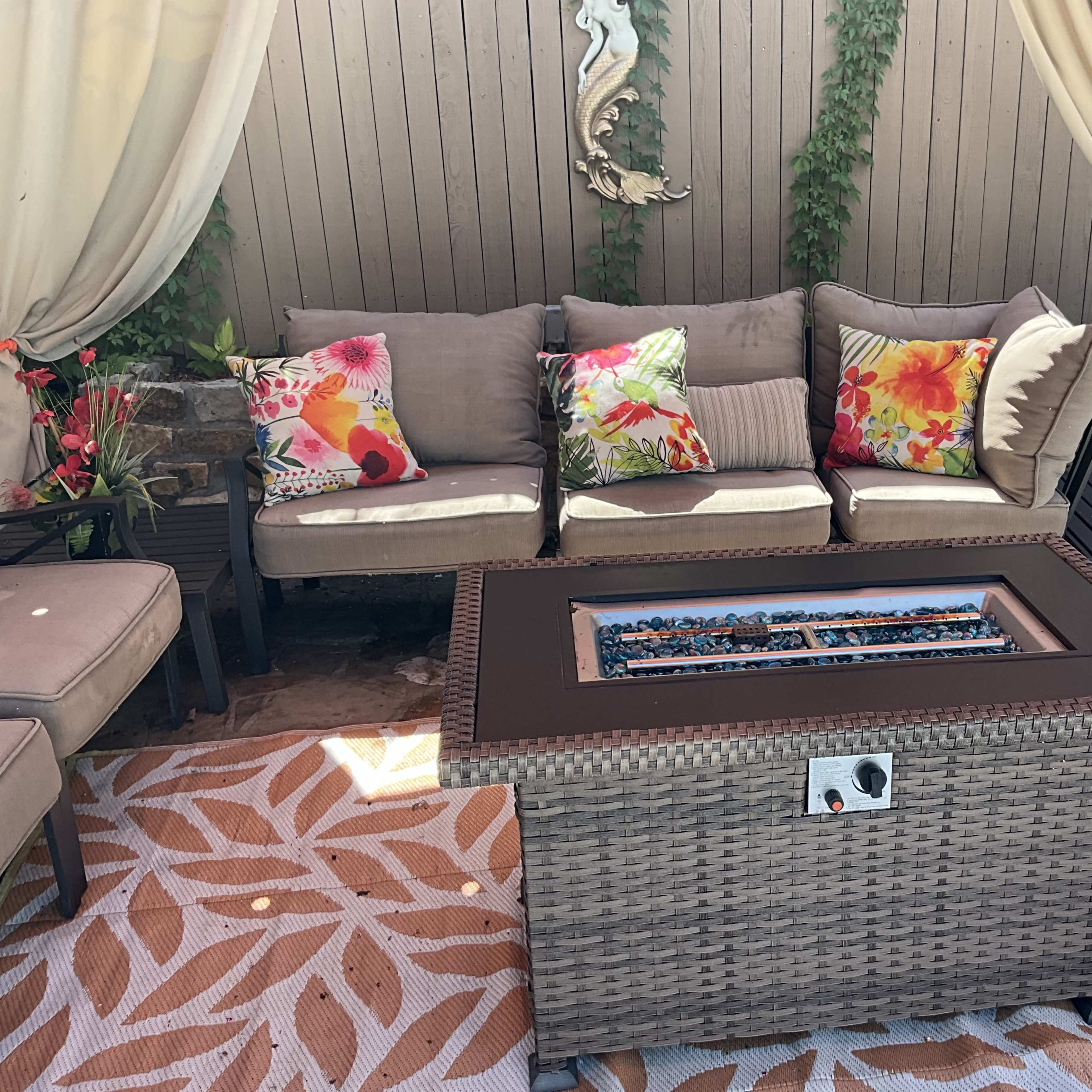 A patio area with a woven coffee table featuring a glass-top fire pit surrounded by a sectional sofa with floral-patterned cushions and draped curtains.