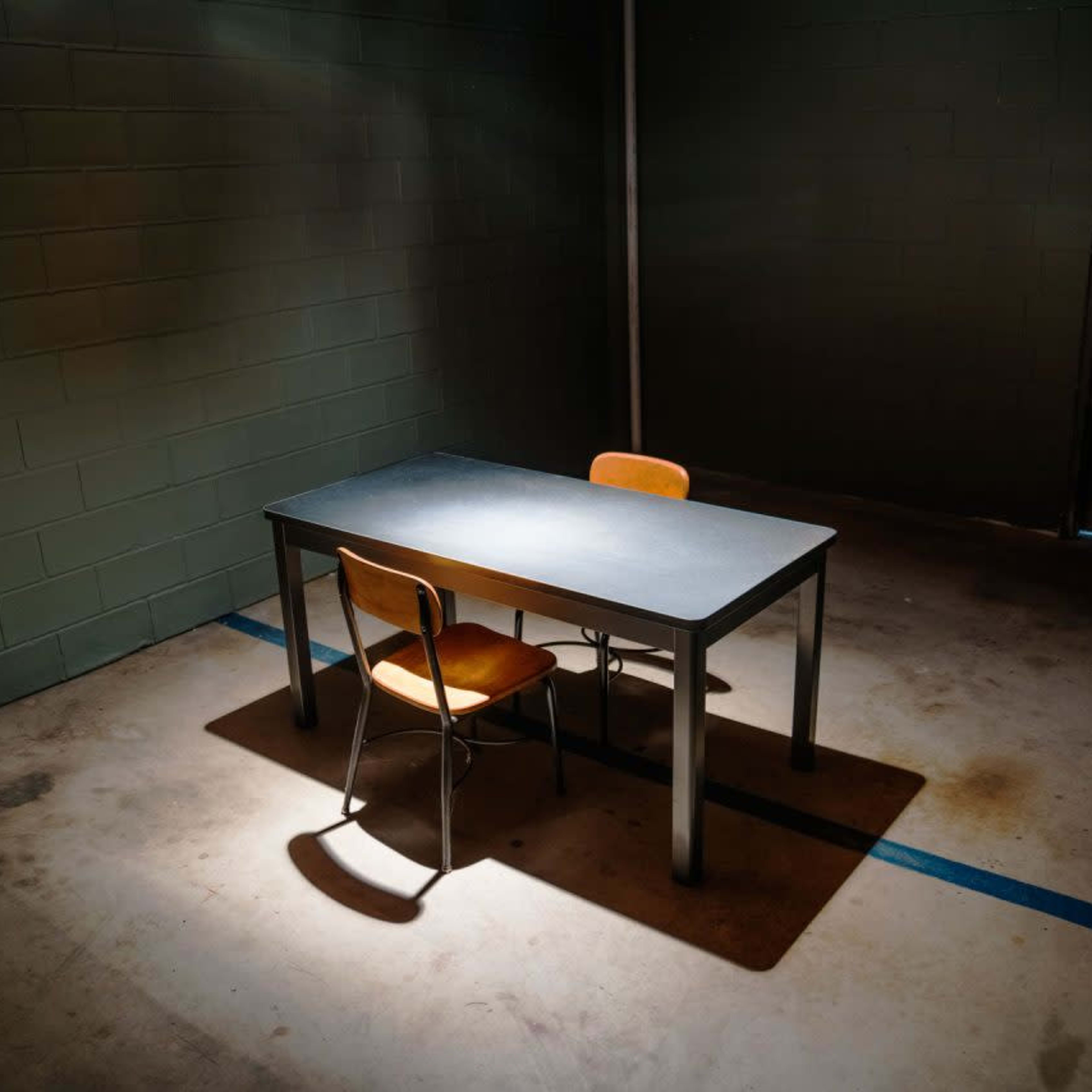 A single table with two chairs is illuminated in a dimly lit room with a concrete floor and a door in the background.