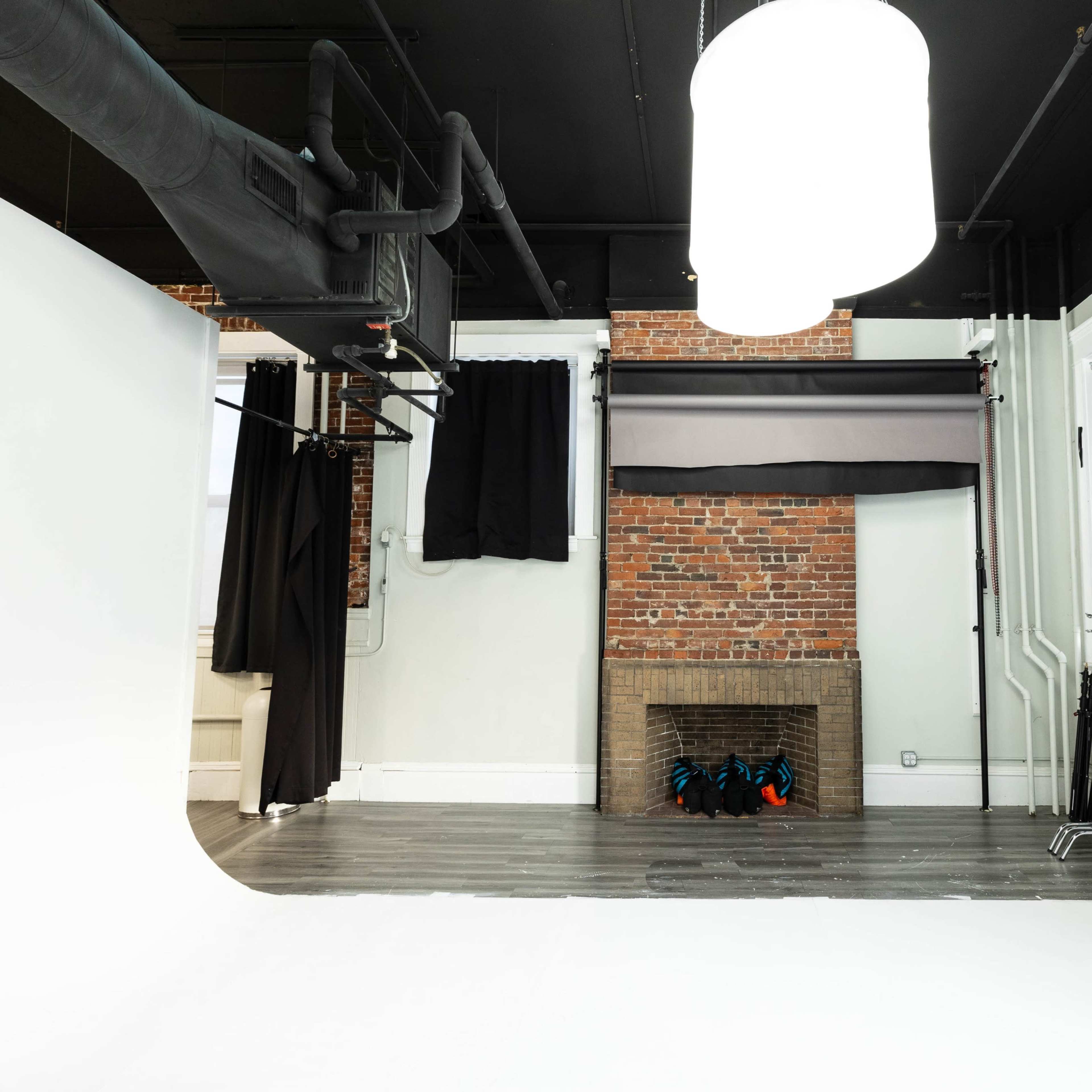 A photography studio featuring a seamless white backdrop, a brick wall with a fireplace, and dark curtains covering windows.