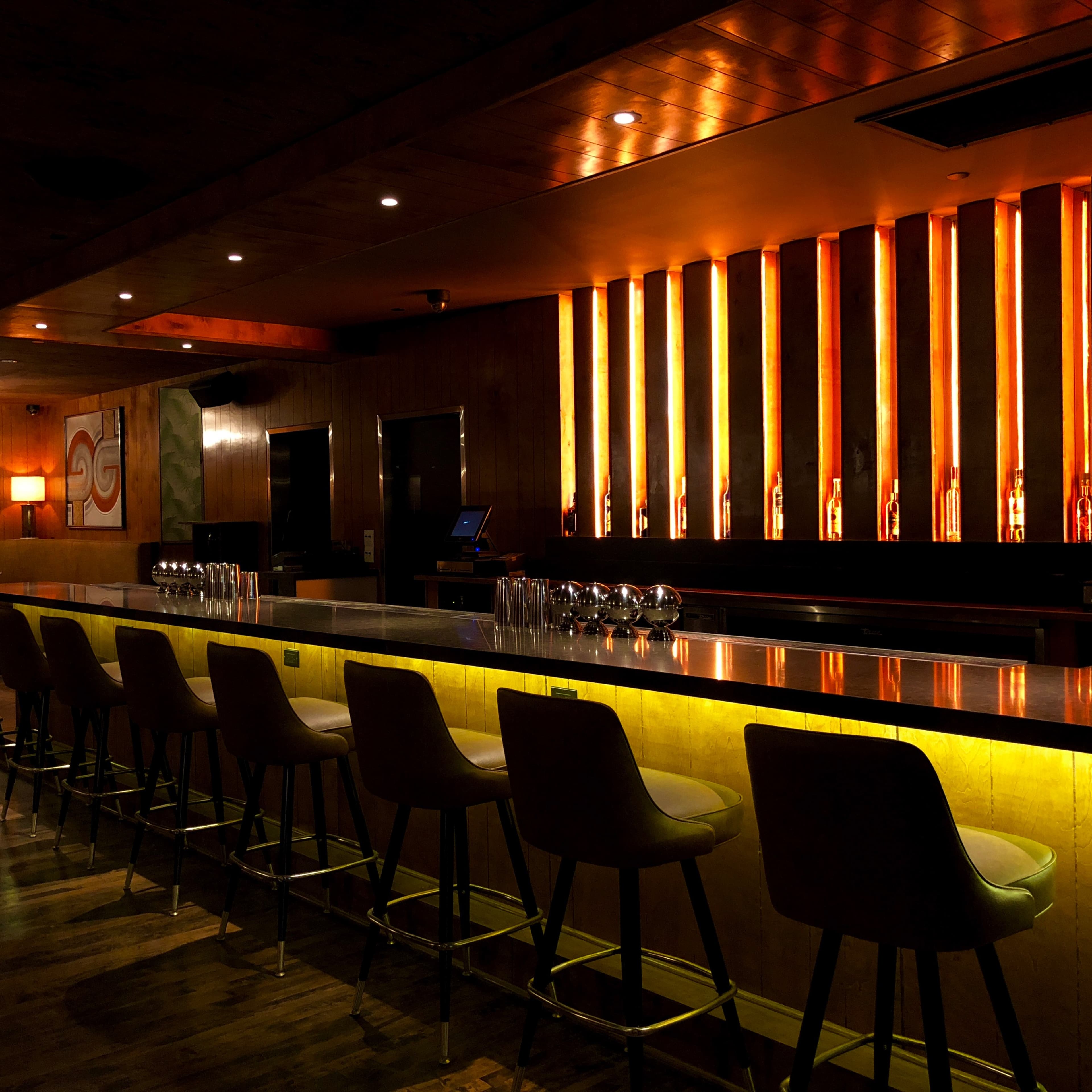 A sleek bar with high stools and illuminated orange vertical panels runs along one side of a dimly lit room.