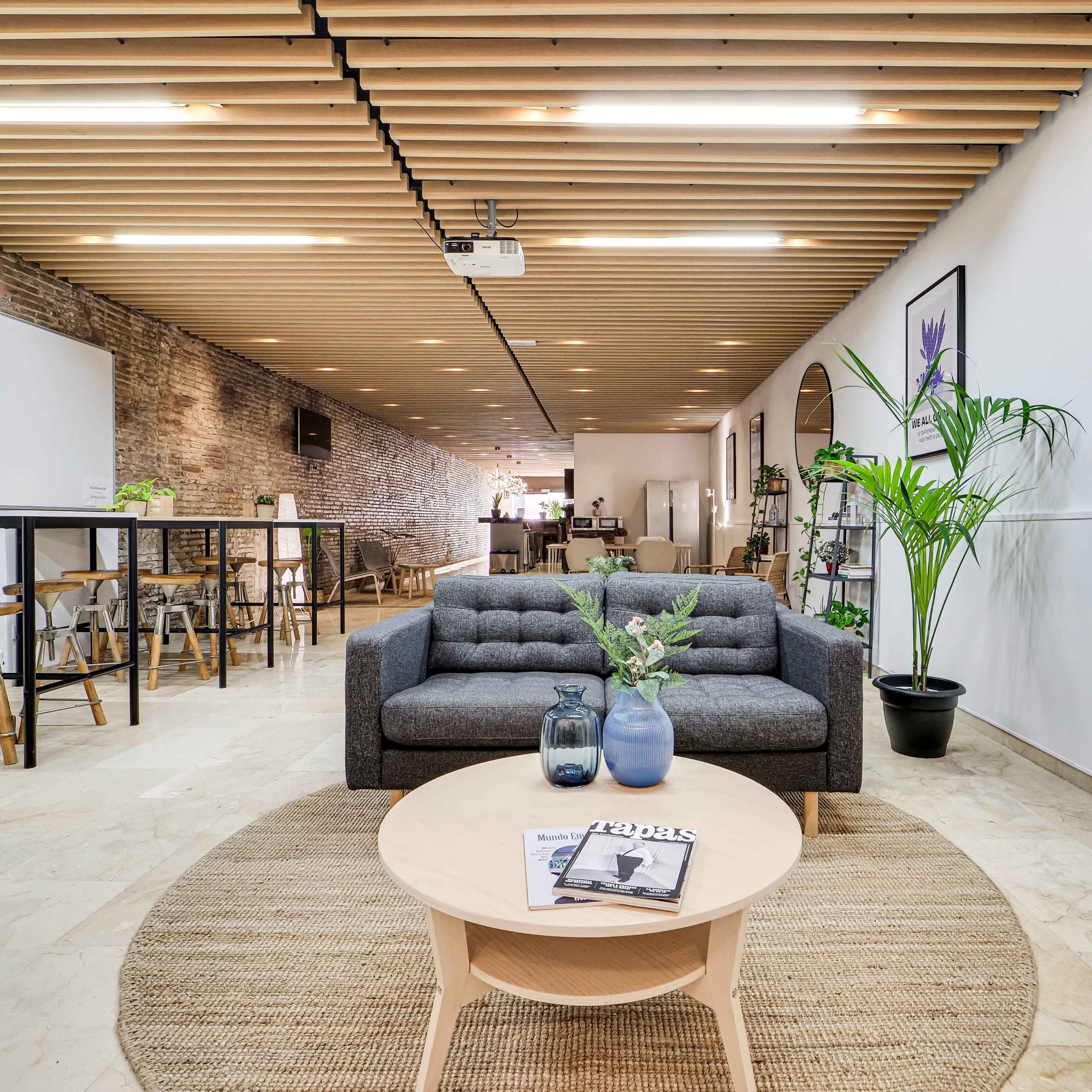 A modern interior space featuring a gray sofa, a round coffee table with a magazine on it, and a plant, set against a backdrop of exposed brick walls and wooden ceiling beams.