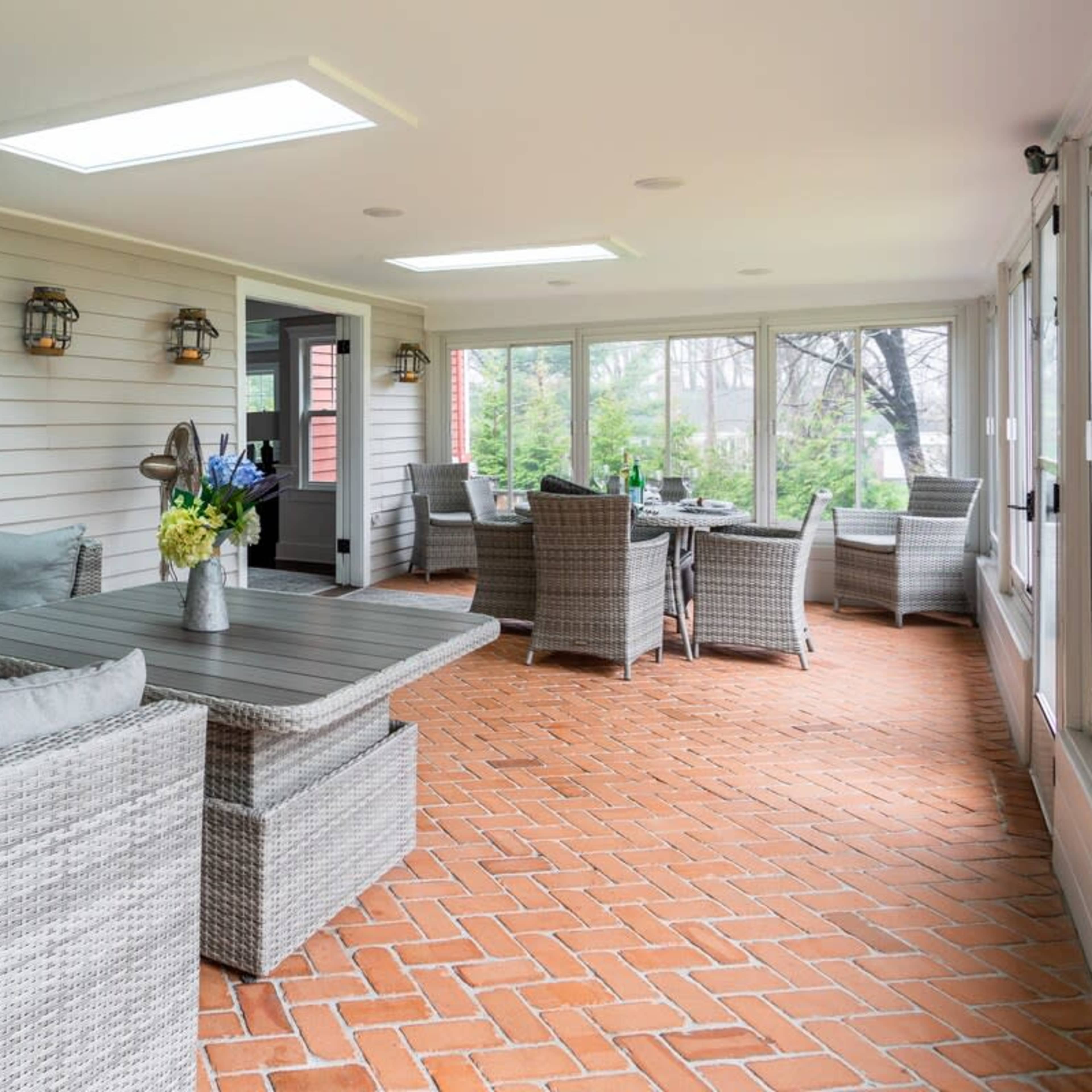 A sunlit enclosed porch with comfortable seating and a table, featuring brick flooring and large windows.