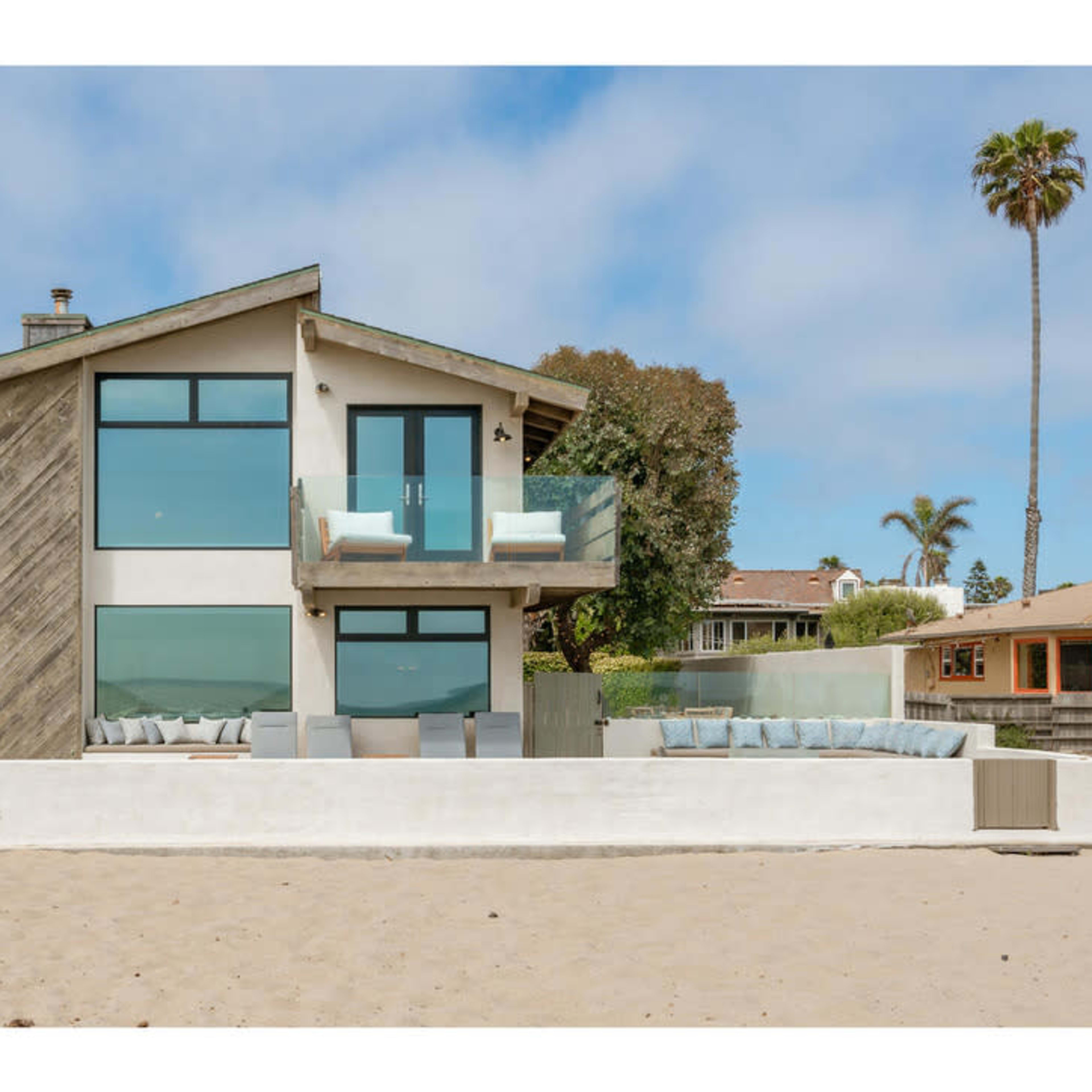 A modern beach house with large windows and a balcony stands along a sandy shore, adjacent to two neighboring homes.