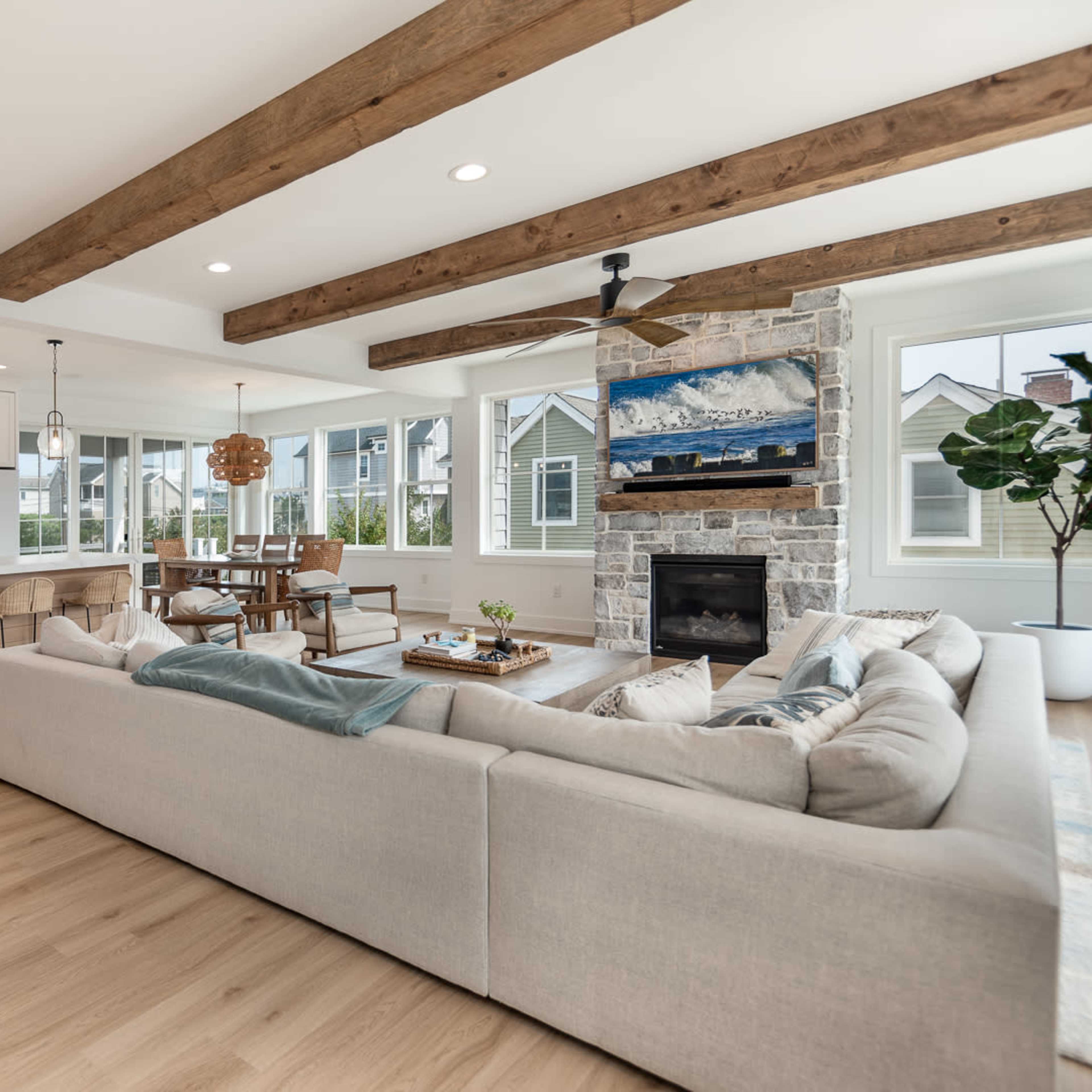 A spacious living area features a sectional sofa, wooden beams on the ceiling, and a stone fireplace, with a dining table and kitchen visible in the background.