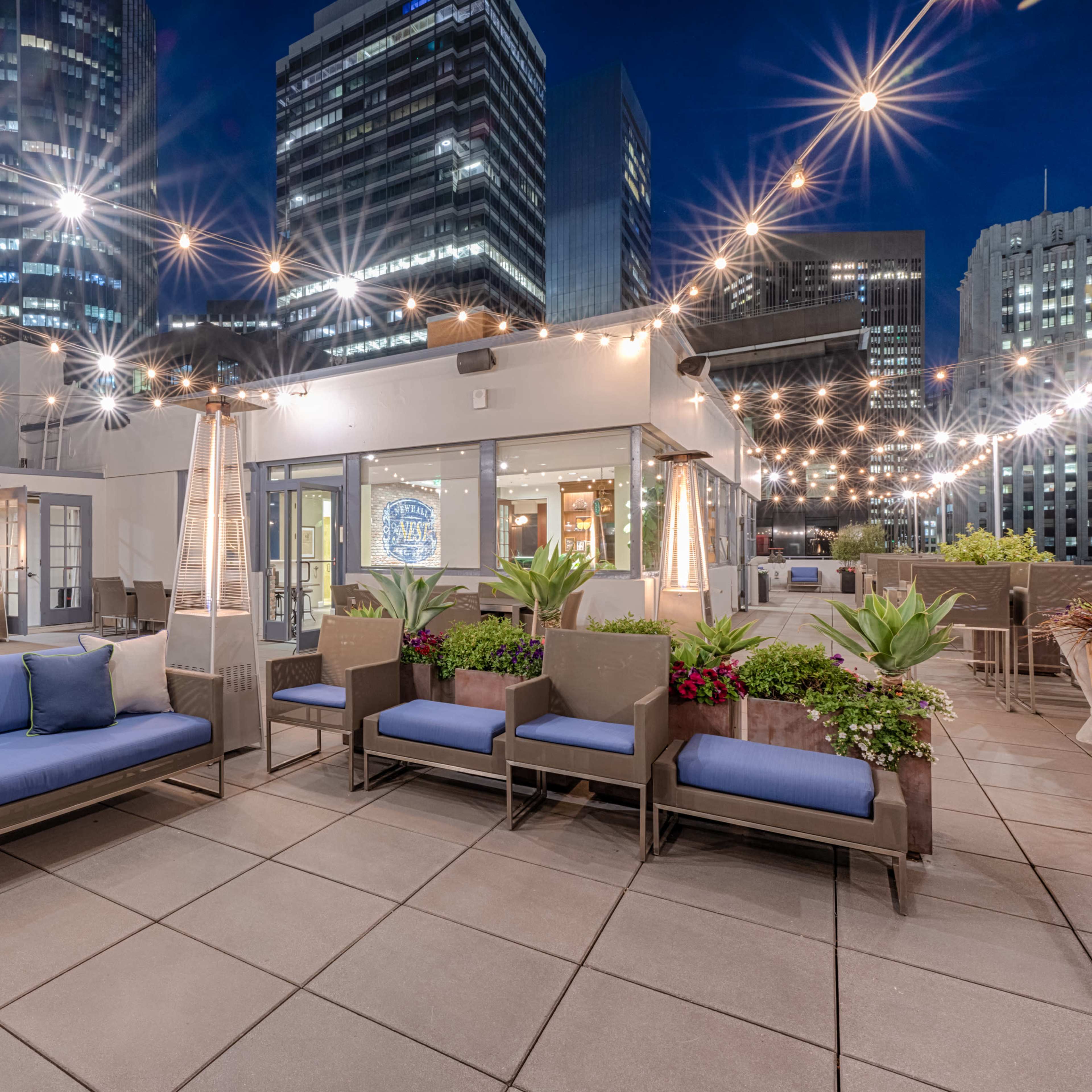 A rooftop lounge area features blue seating, potted plants, and string lights against a backdrop of city skyscrapers at night.