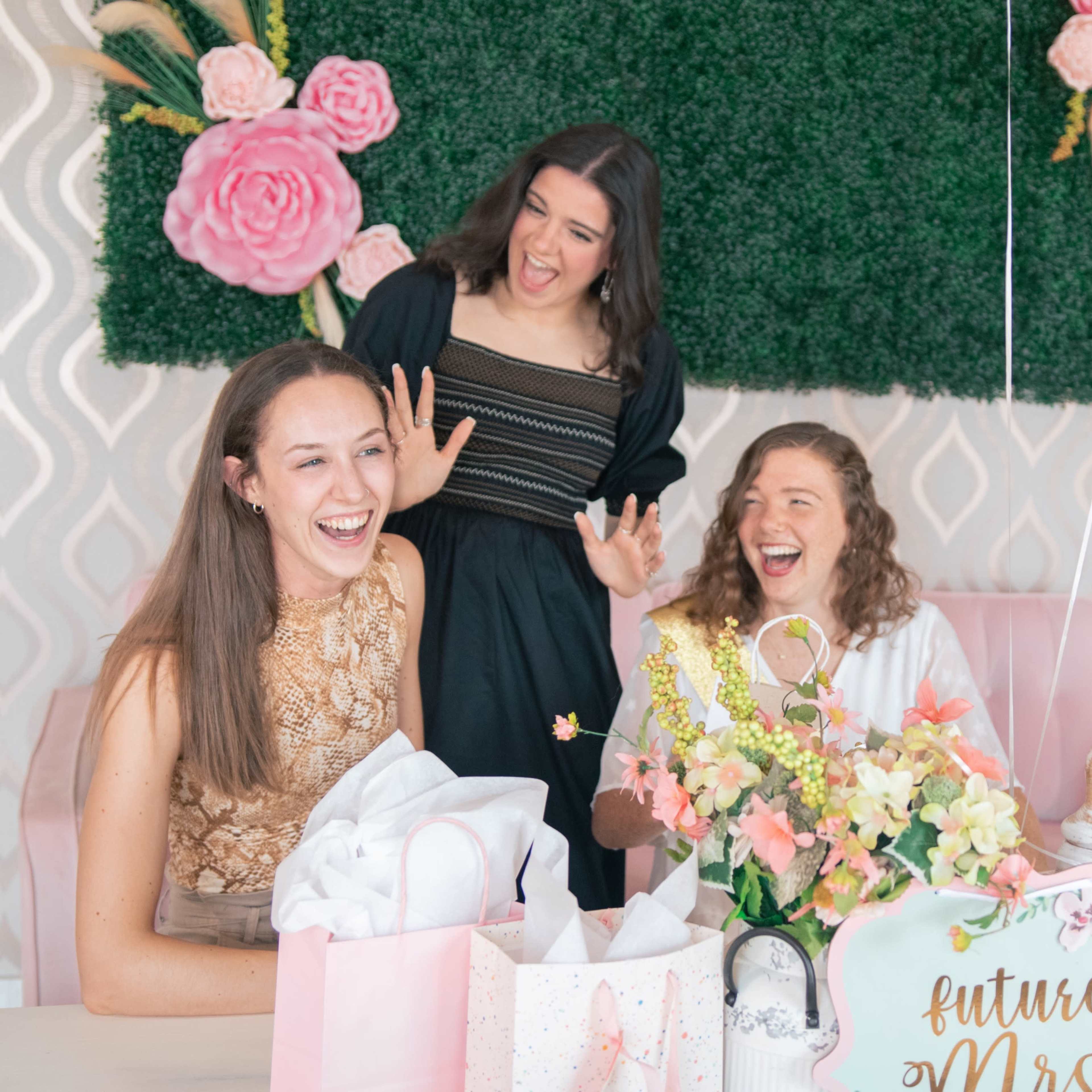 Four women sit at a table decorated with flowers and gifts, laughing and engaging joyfully during a celebratory event.