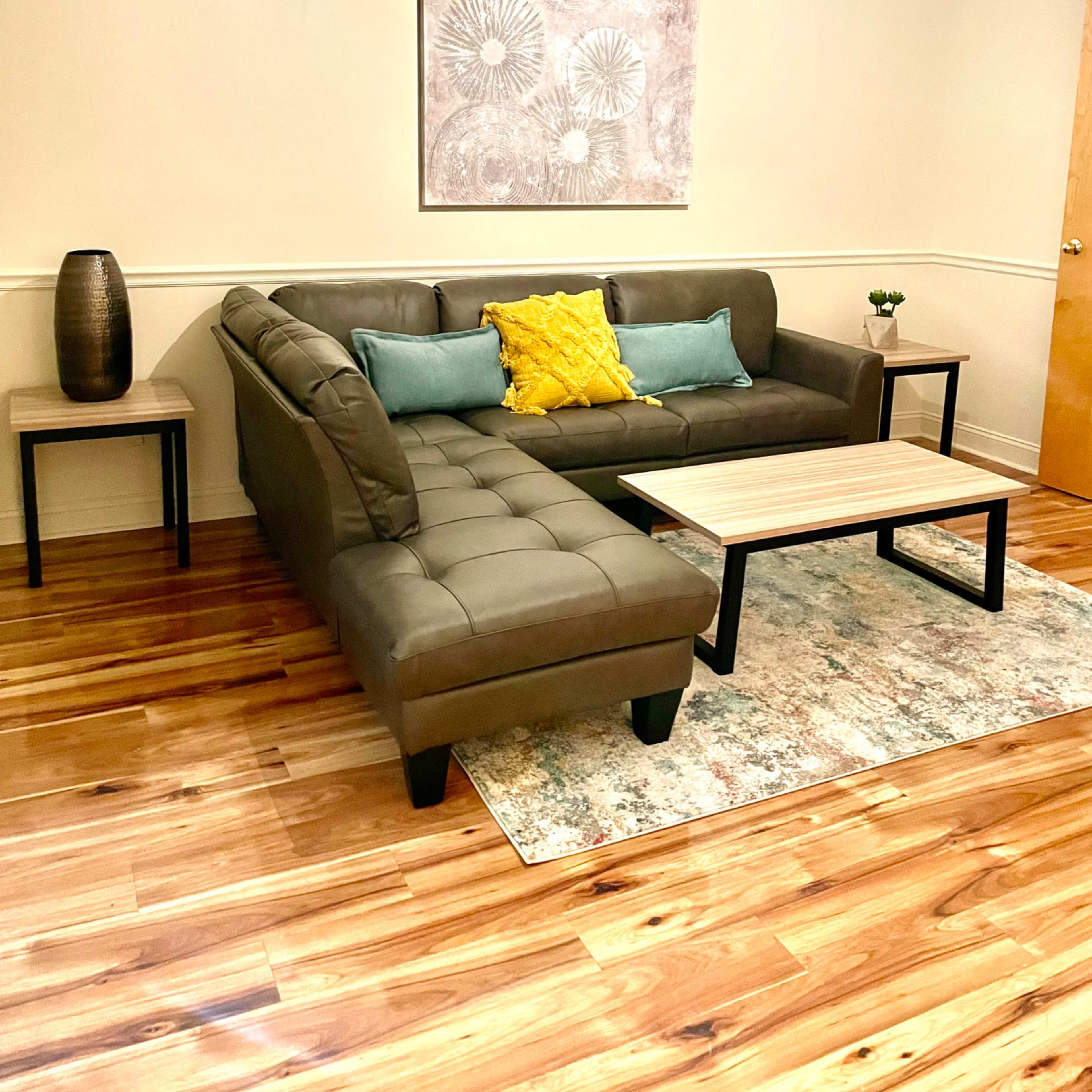 A modern living room features a gray sectional sofa with colorful cushions, a small wooden coffee table, and a decorative vase on a side table, all set on a wooden floor.