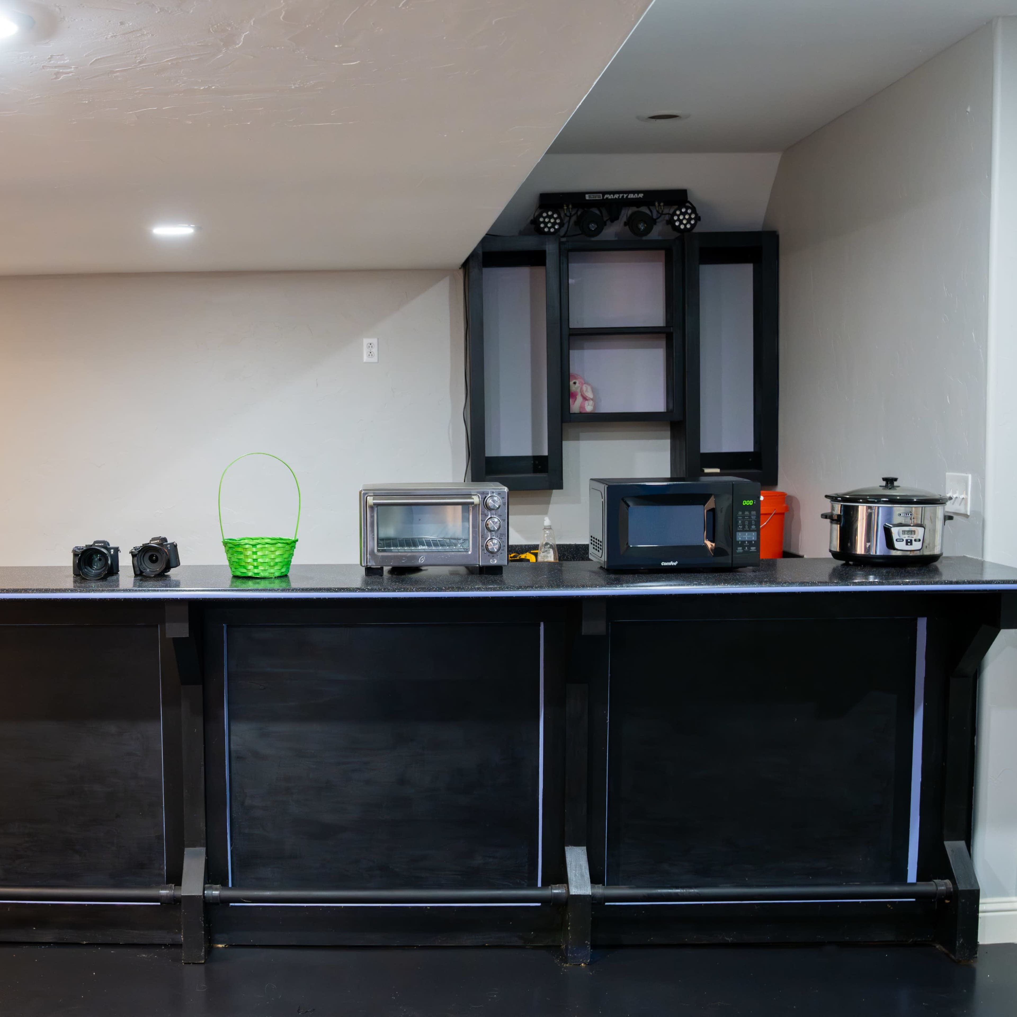 A modern bar area features a black countertop with several appliances, including a microwave, slow cooker, and camera equipment, against a neutral-colored wall.