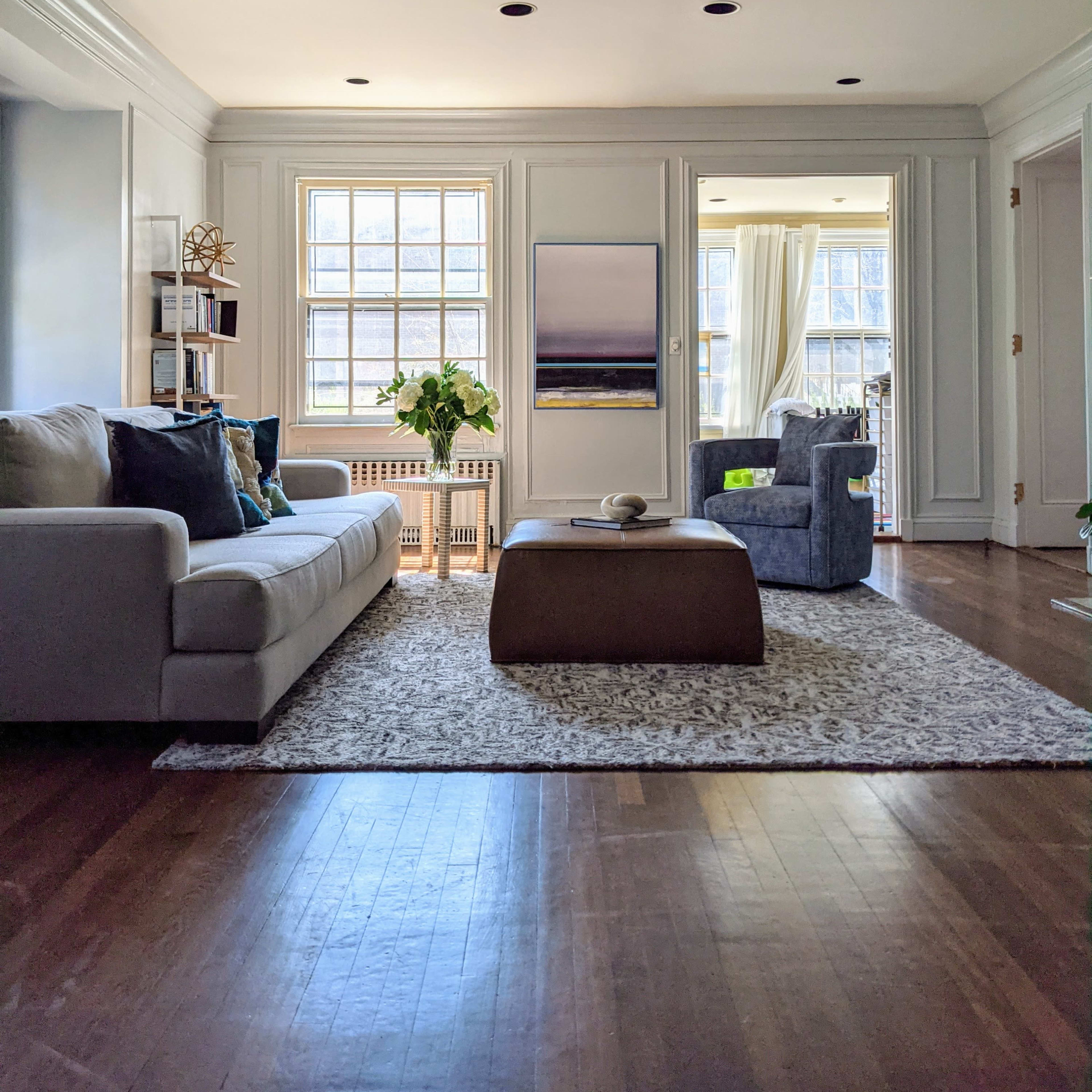 The living room features a light-colored sofa, a round coffee table, and a cozy armchair, with large windows providing ample natural light.