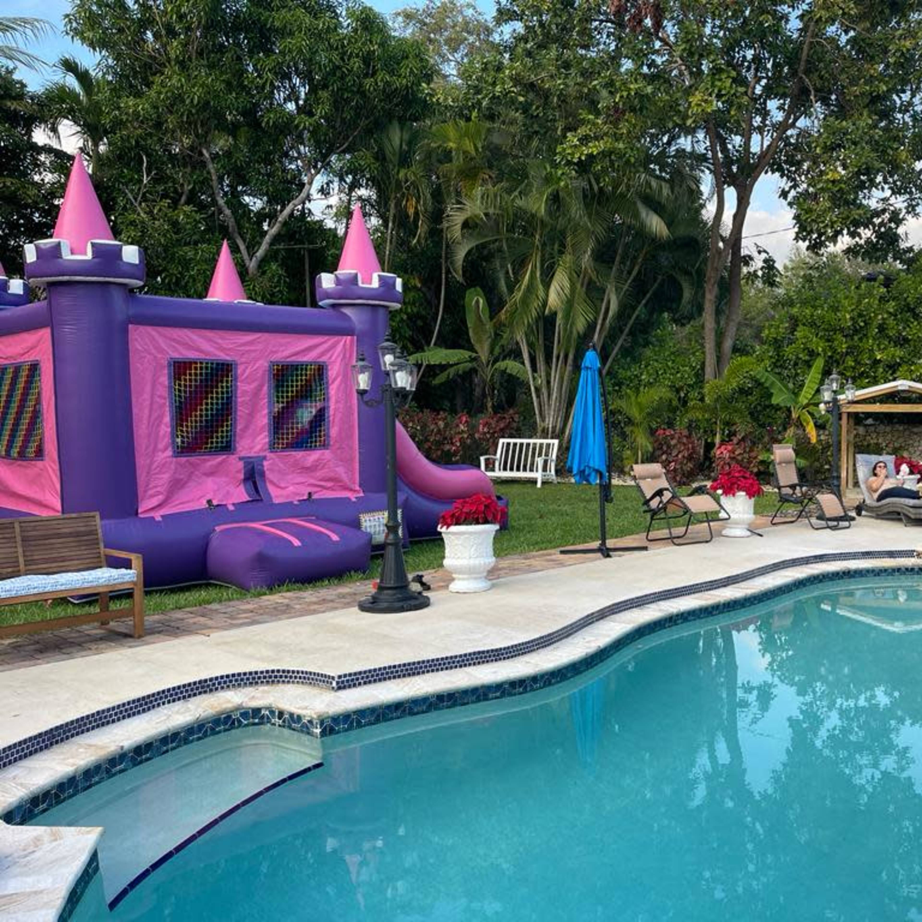 The scene depicts a backyard pool area featuring a pink inflatable bouncy castle, lounge chairs, and potted plants, surrounded by lush greenery.