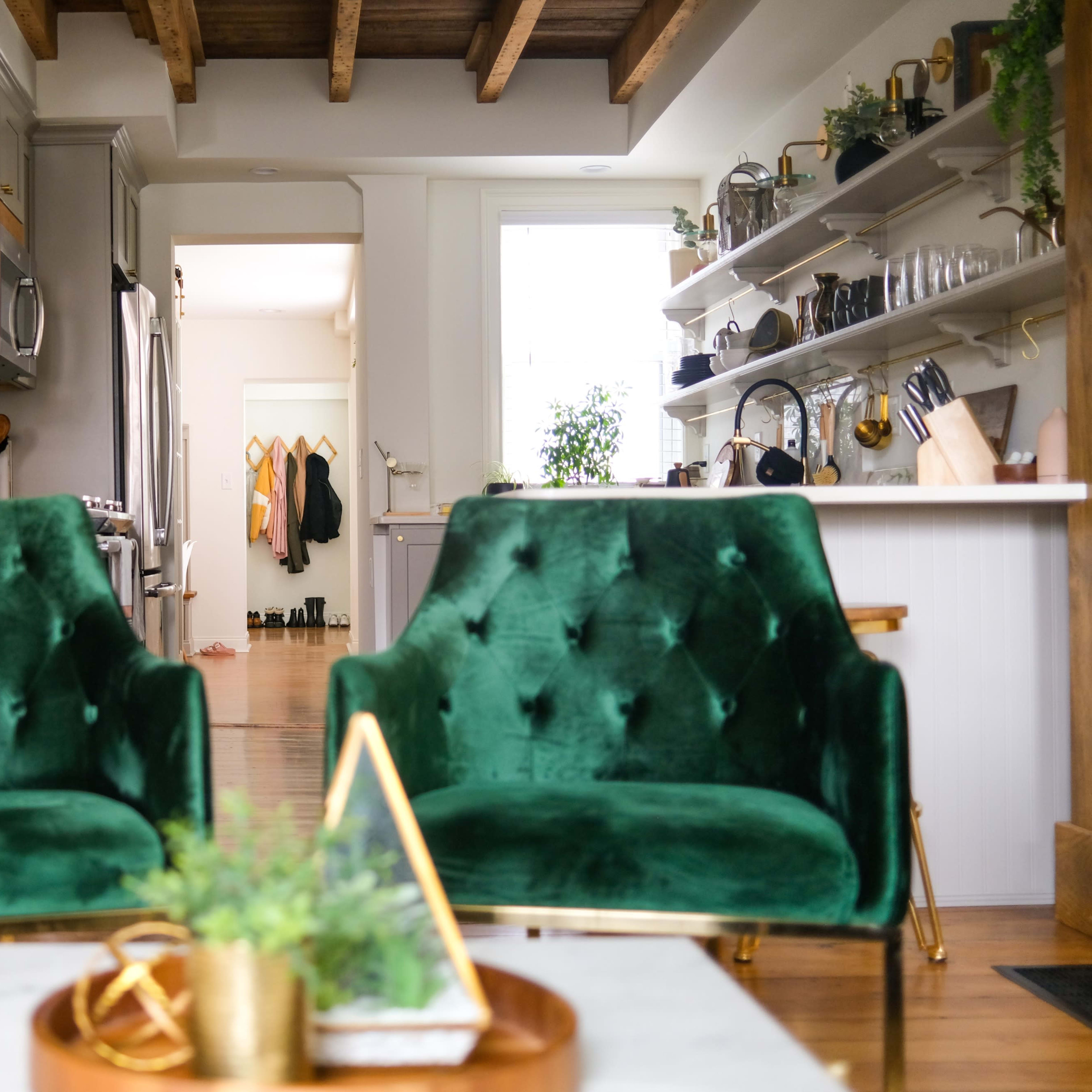 Two green velvet chairs facing a dining area with an open kitchen, shelves displaying kitchenware, and a staircase in the background.