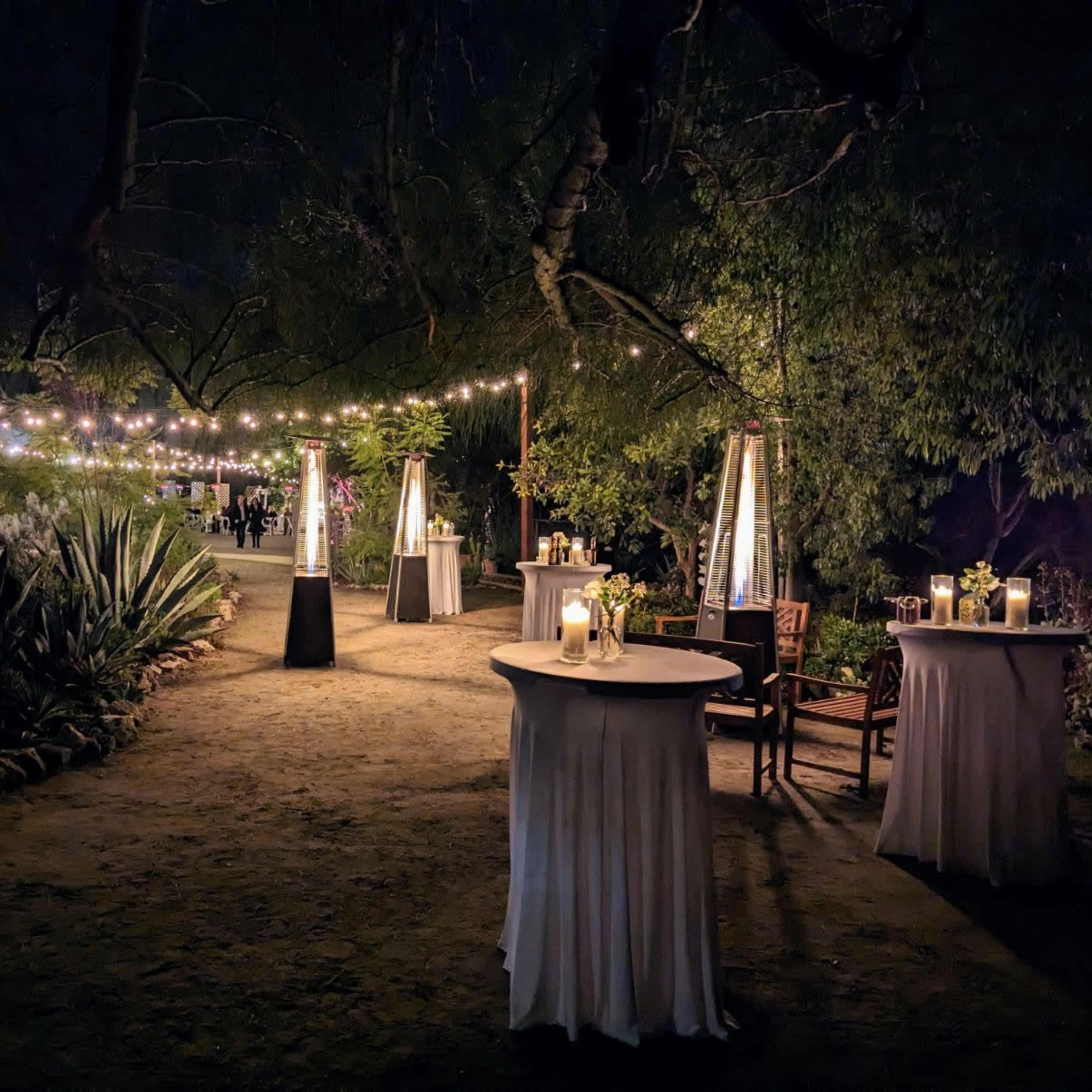 A beautifully lit outdoor event space with round tables and lanterns under strings of lights.