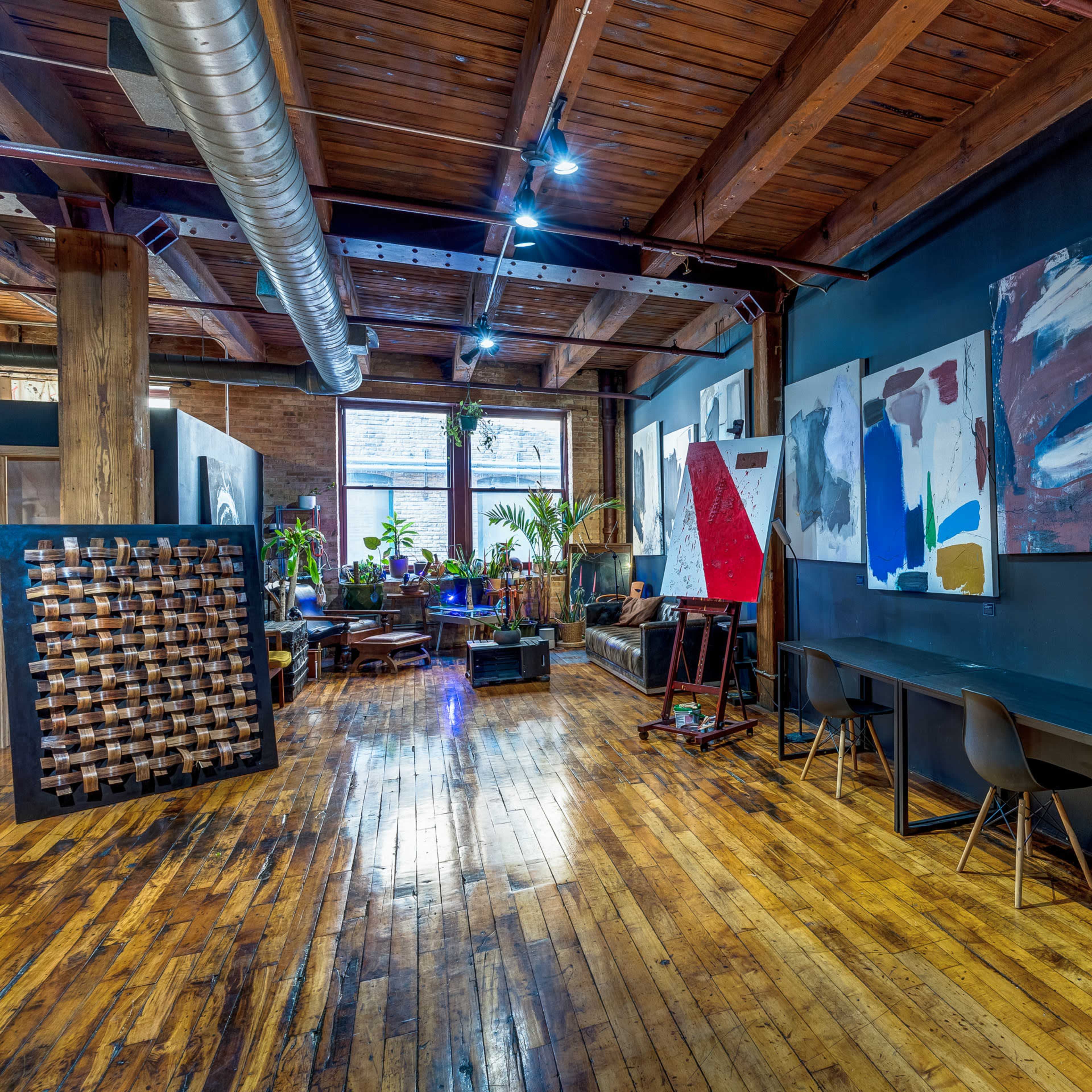 A spacious, industrial-style room with wooden floors, dark walls, and various large paintings hanging, along with a few seating areas and plants near a window.
