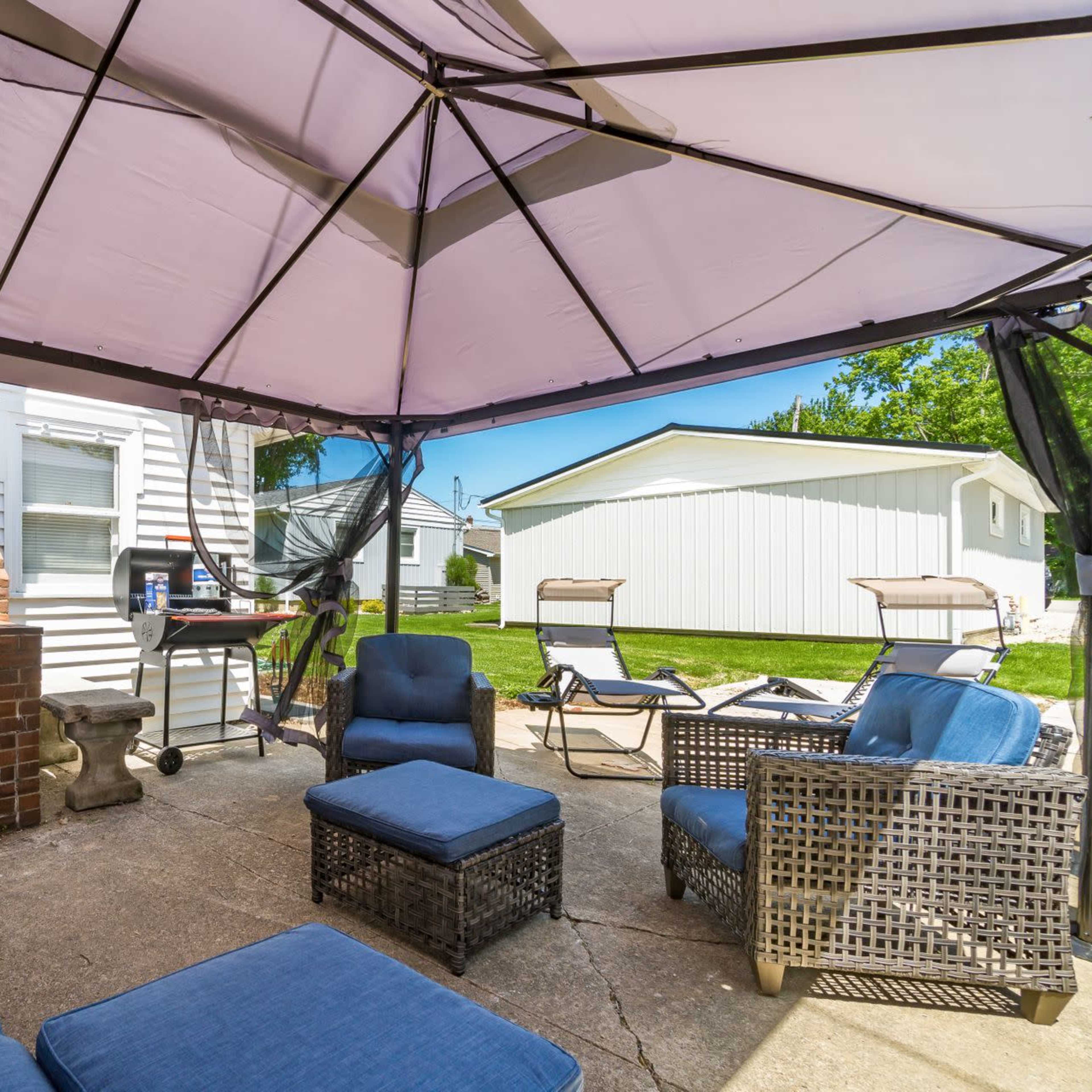 A shaded outdoor seating area with blue furniture, a brick barbecue grill, and a view of a lawn and nearby structures.