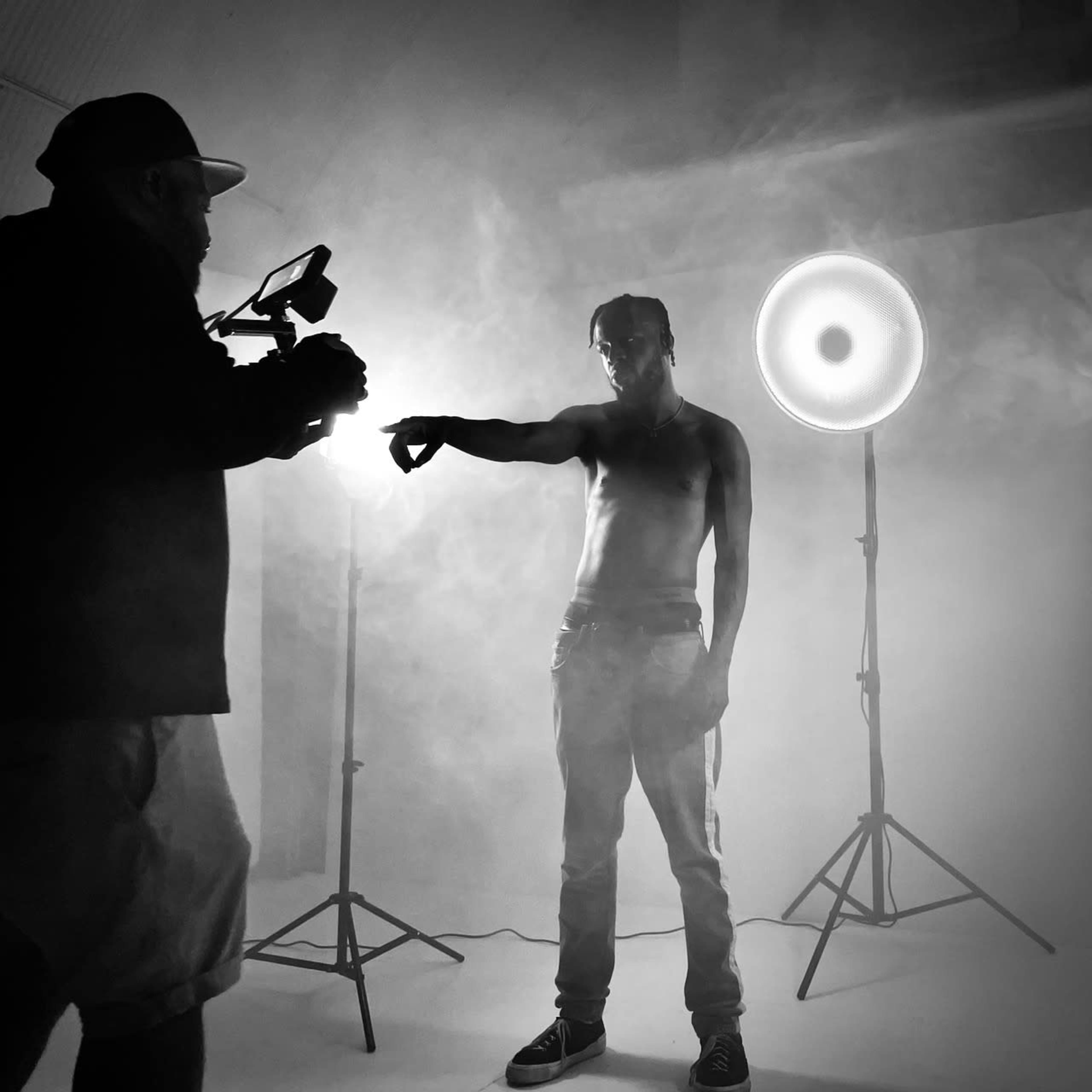 A shirtless individual poses dramatically in a smoke-filled studio while a person behind a camera captures the moment.