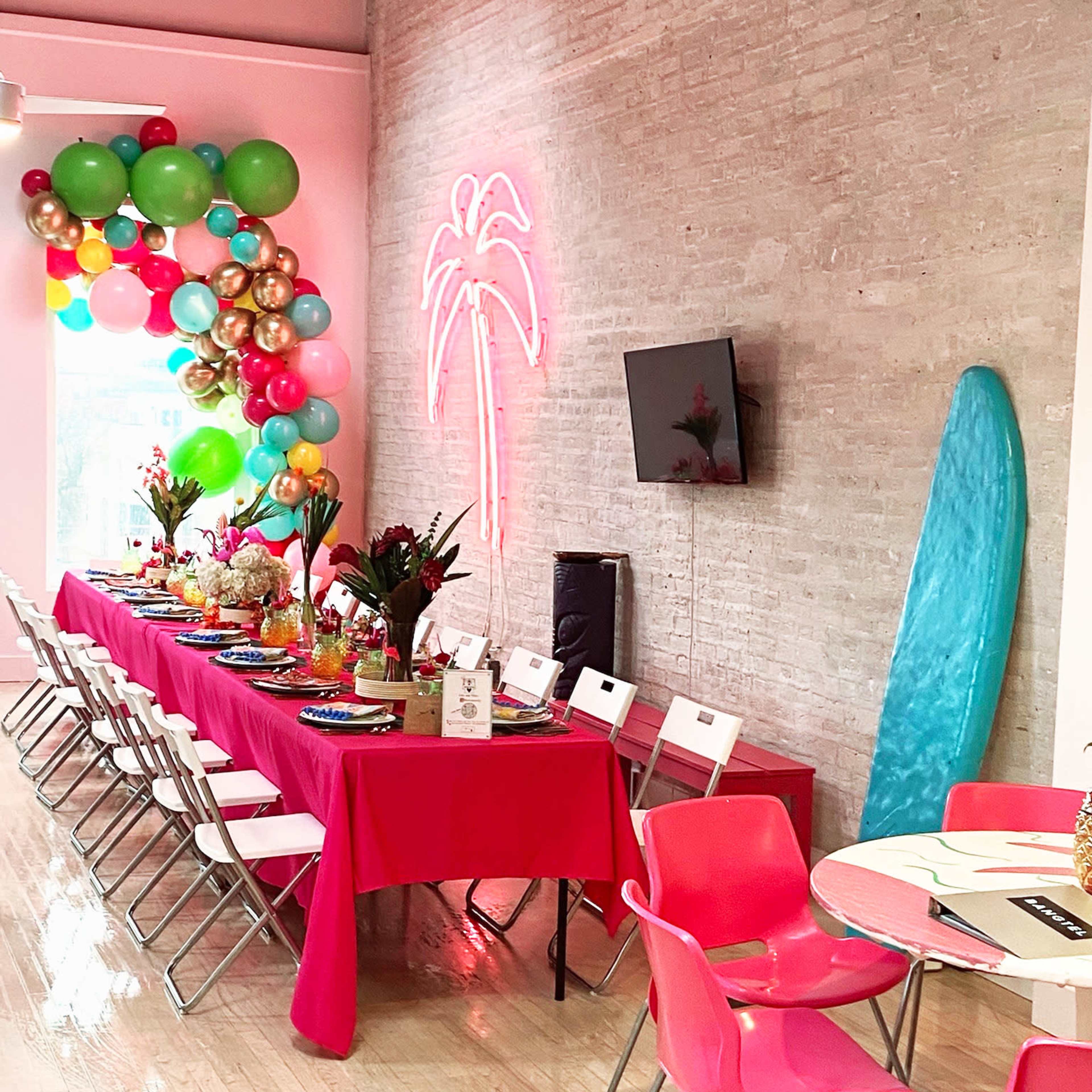 A vibrant party setting features a long table adorned with colorful decorations, surrounded by chairs, with balloons and a neon palm tree sign accentuating the room.