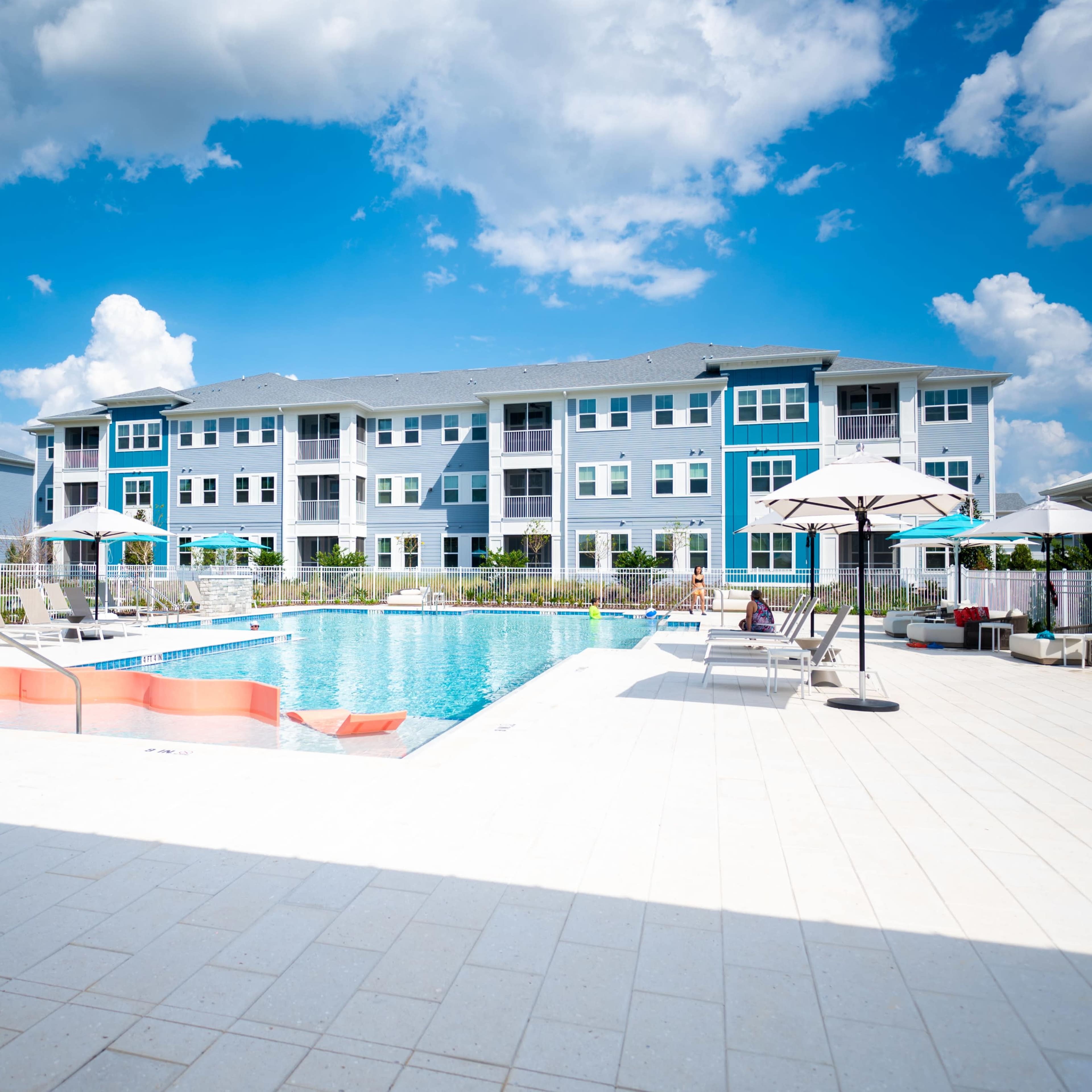 A modern apartment complex with a swimming pool and lounge area under clear blue skies.