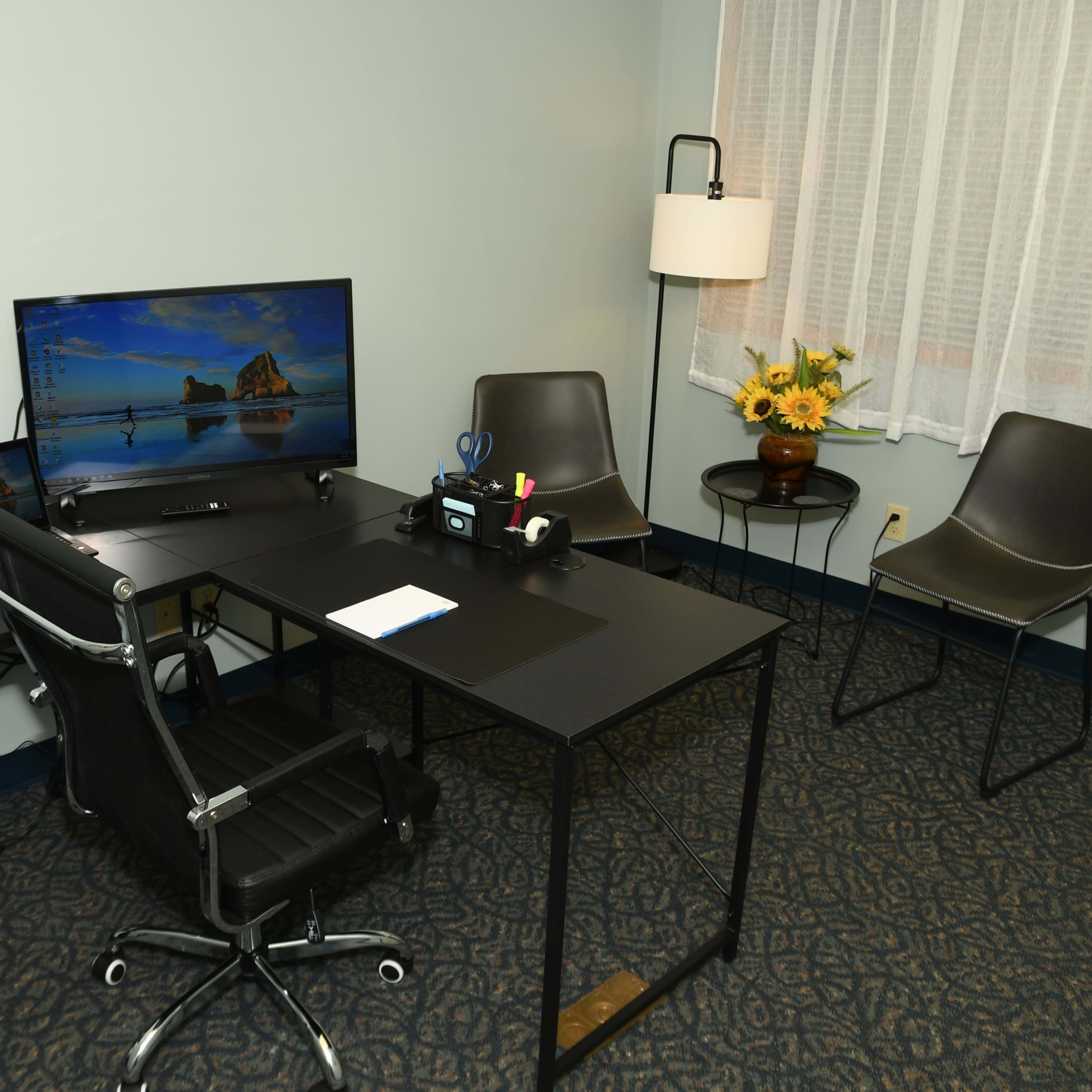 A modern office space equipped with a desk, two monitors, a black office chair, and a small seating area with two chairs and a lamp.