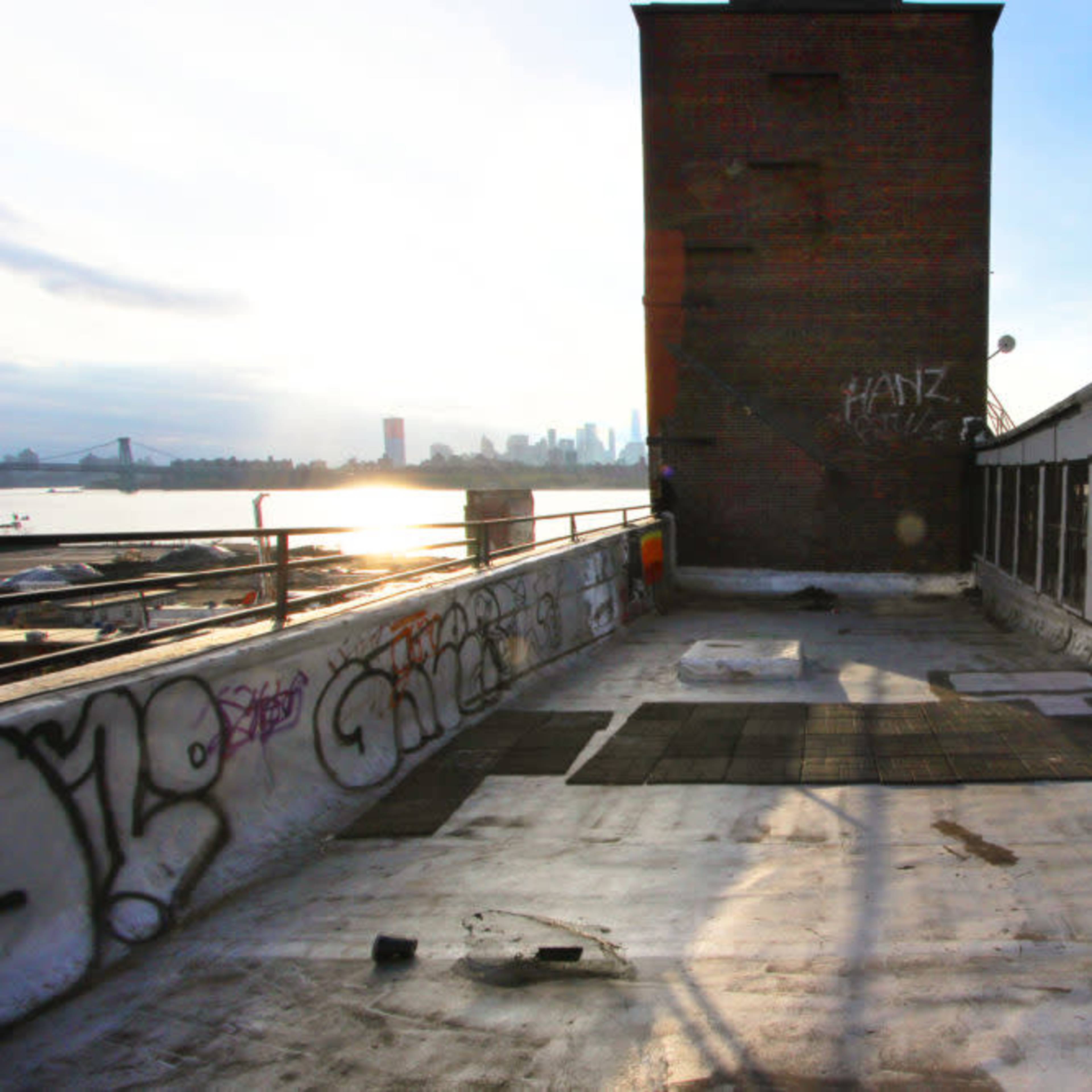 A rooftop with a brick structure and graffiti, overlooking a river and a city skyline in the background.