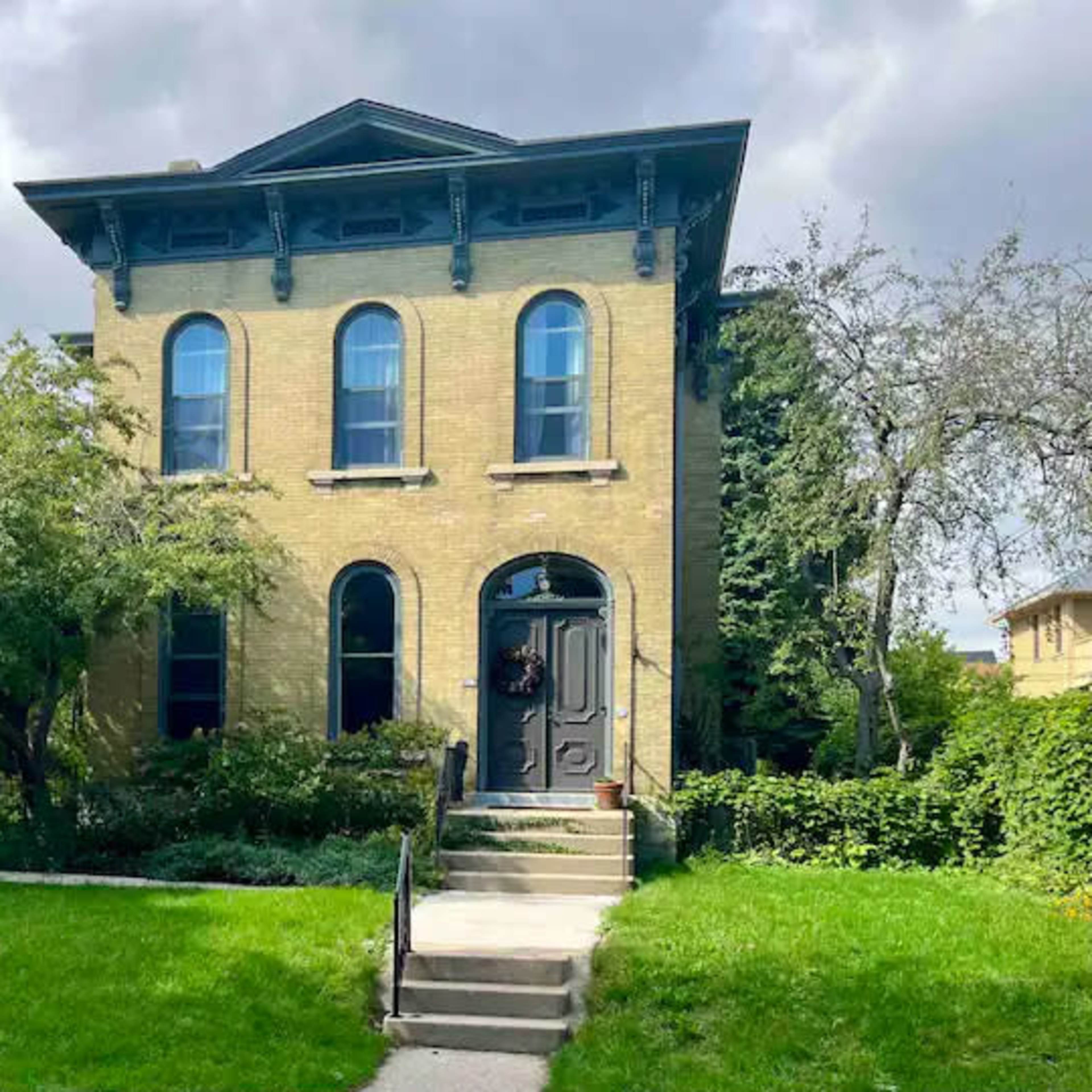 A historic two-story brick house features arched windows and a central front door, surrounded by greenery and a well-maintained lawn.