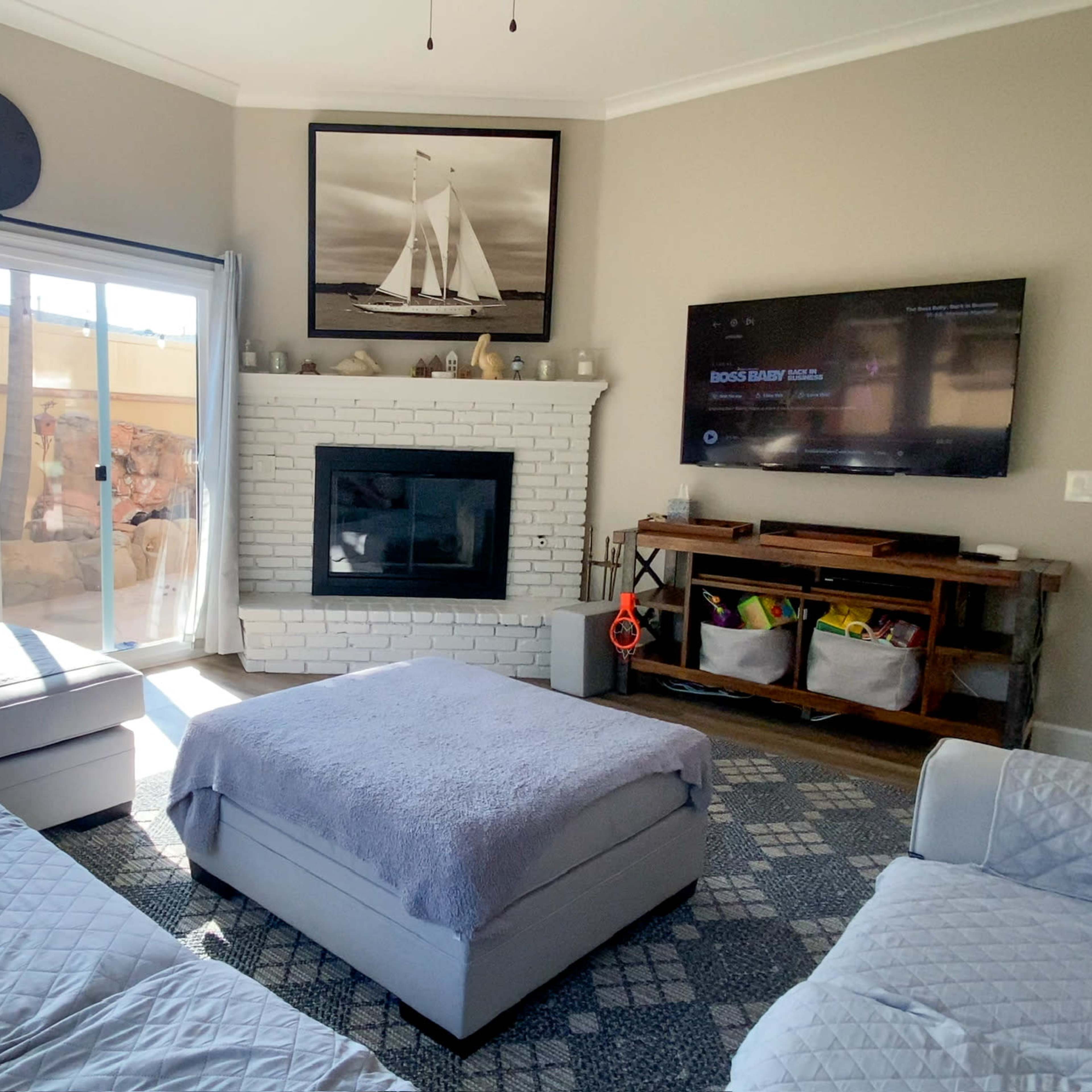 The living room features a white brick fireplace under a wall-mounted TV, with a large window overlooking an outdoor space, and seating arranged around a light-colored ottoman.