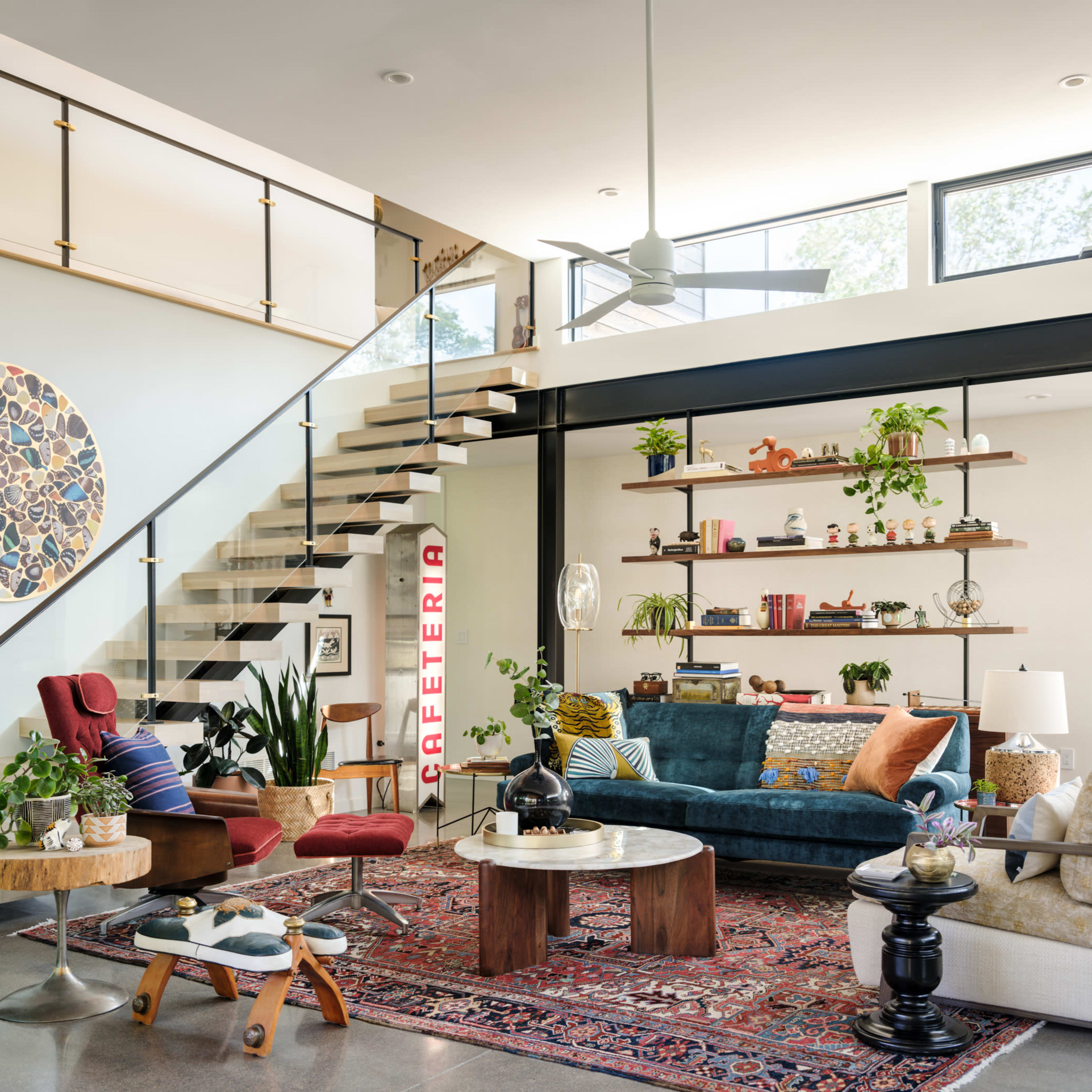 A modern living room with a staircase, various plants, a colorful rug, and a mix of seating options arranged around a coffee table.