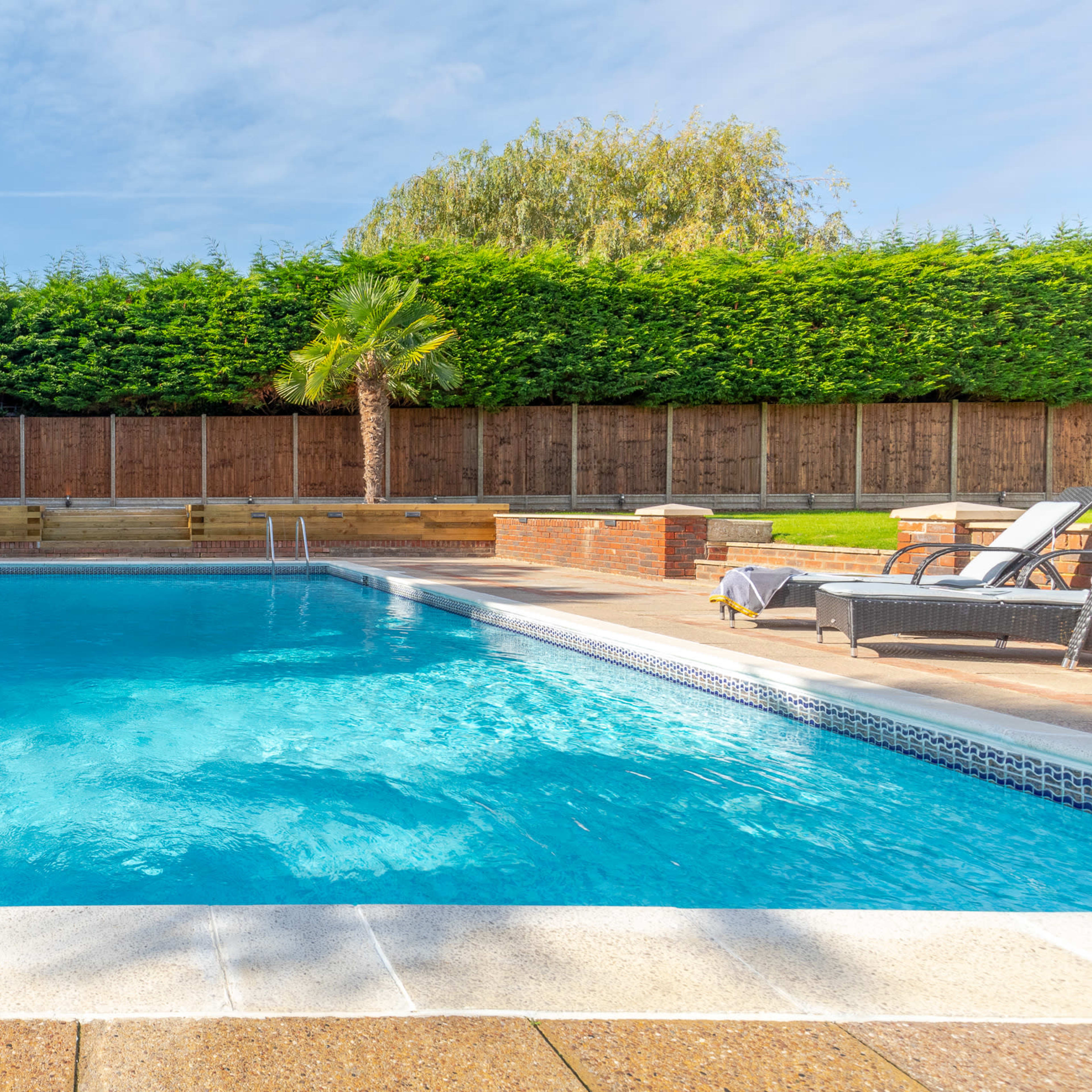 A clear swimming pool surrounded by stone decking, palm trees, and wooden fencing.
