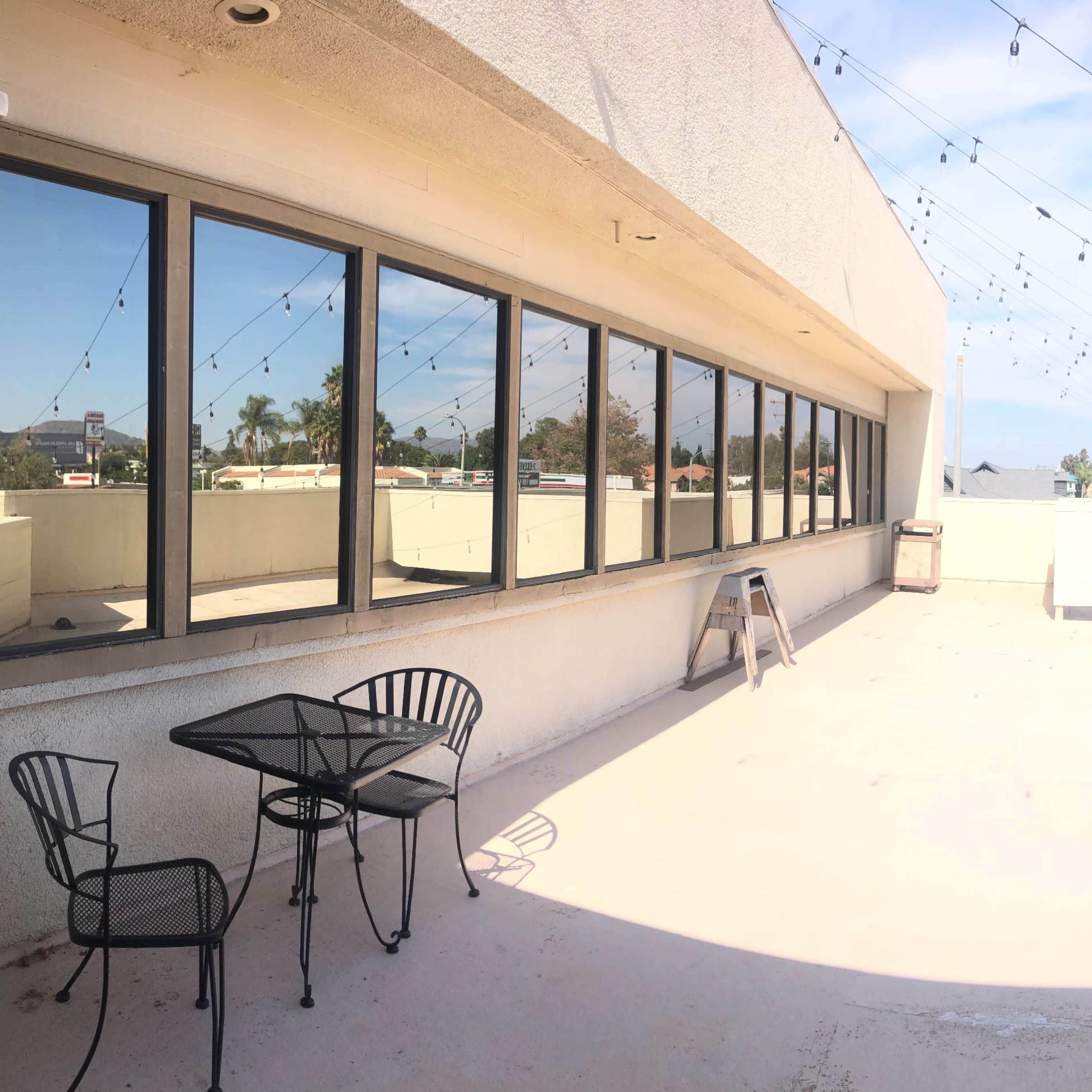 A panoramic view of a rooftop terrace featuring a small table and chairs, with string lights overhead and a clear sky in the background.