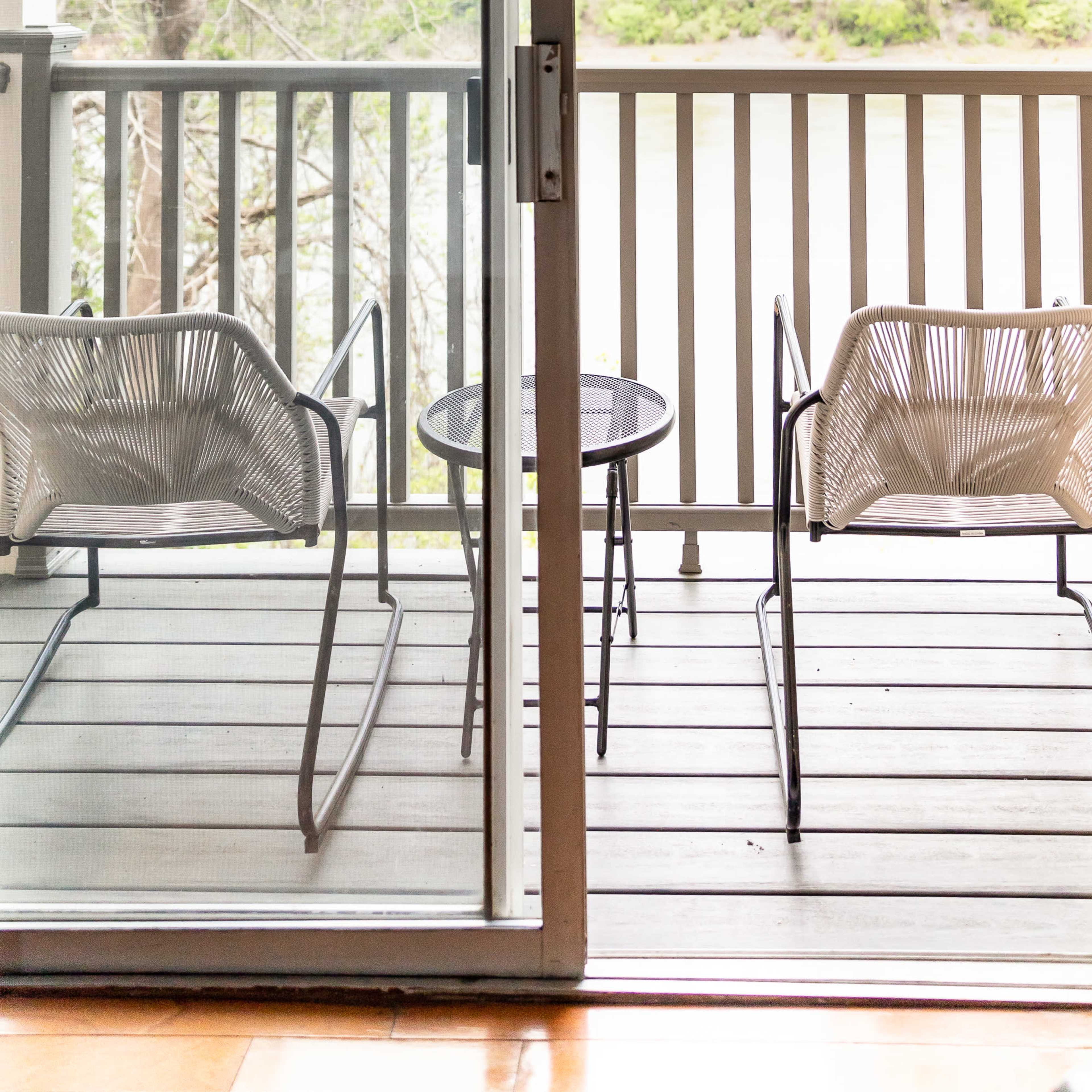 A balcony features two chairs and a small table, overlooking a body of water and greenery.