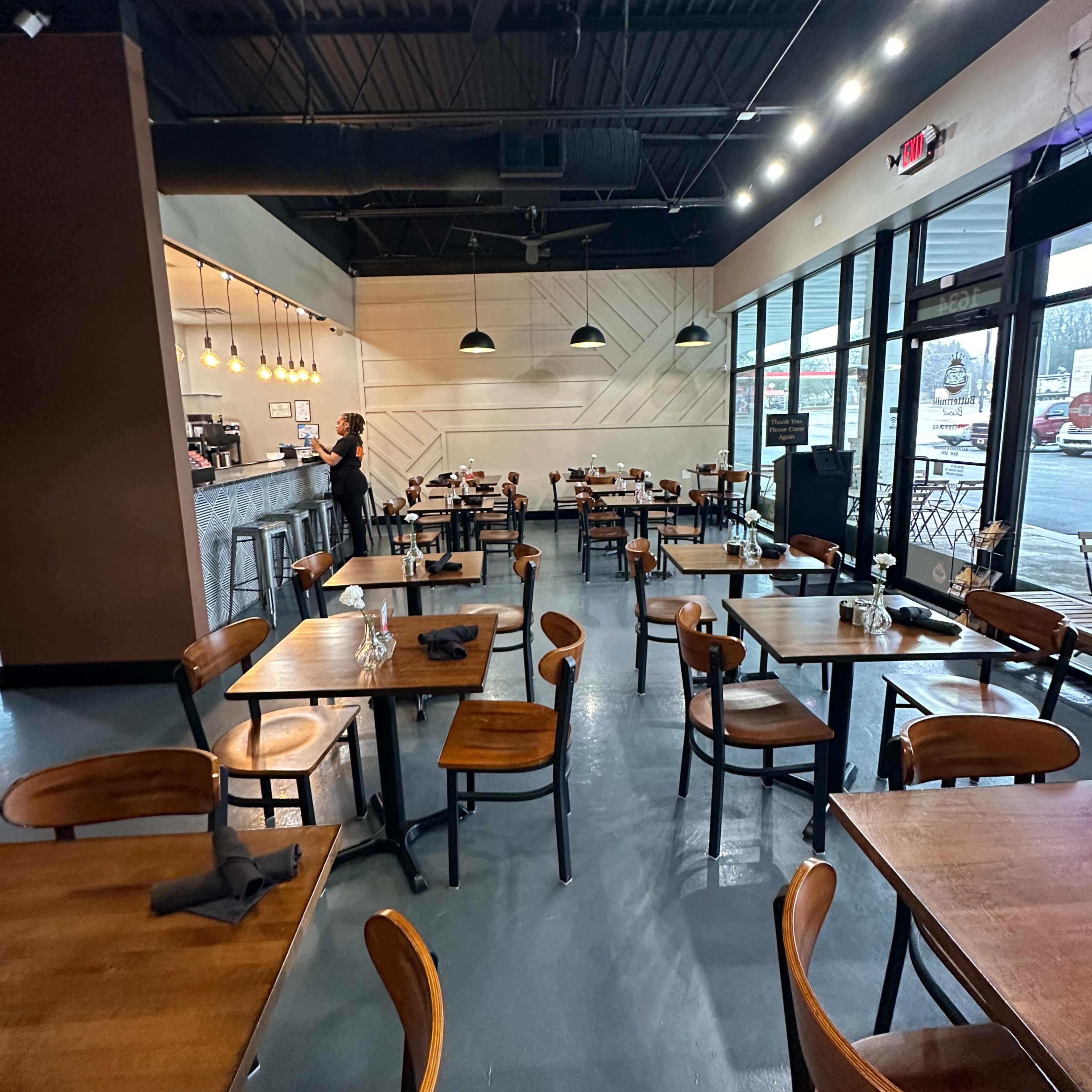 A spacious restaurant interior with wooden tables and chairs arranged neatly, and a staff member standing near the counter.